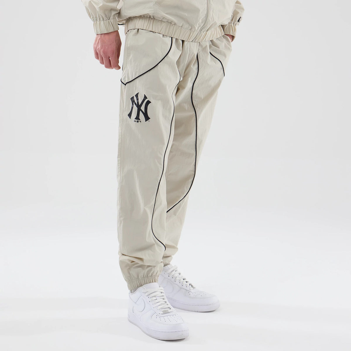 New Era New York Yankees MLB Piping Track Cream Track Pants, front view on a model showing the embroidered NY logo on the left thigh, black piping along the seams, and elasticated cuffs above white trainers.