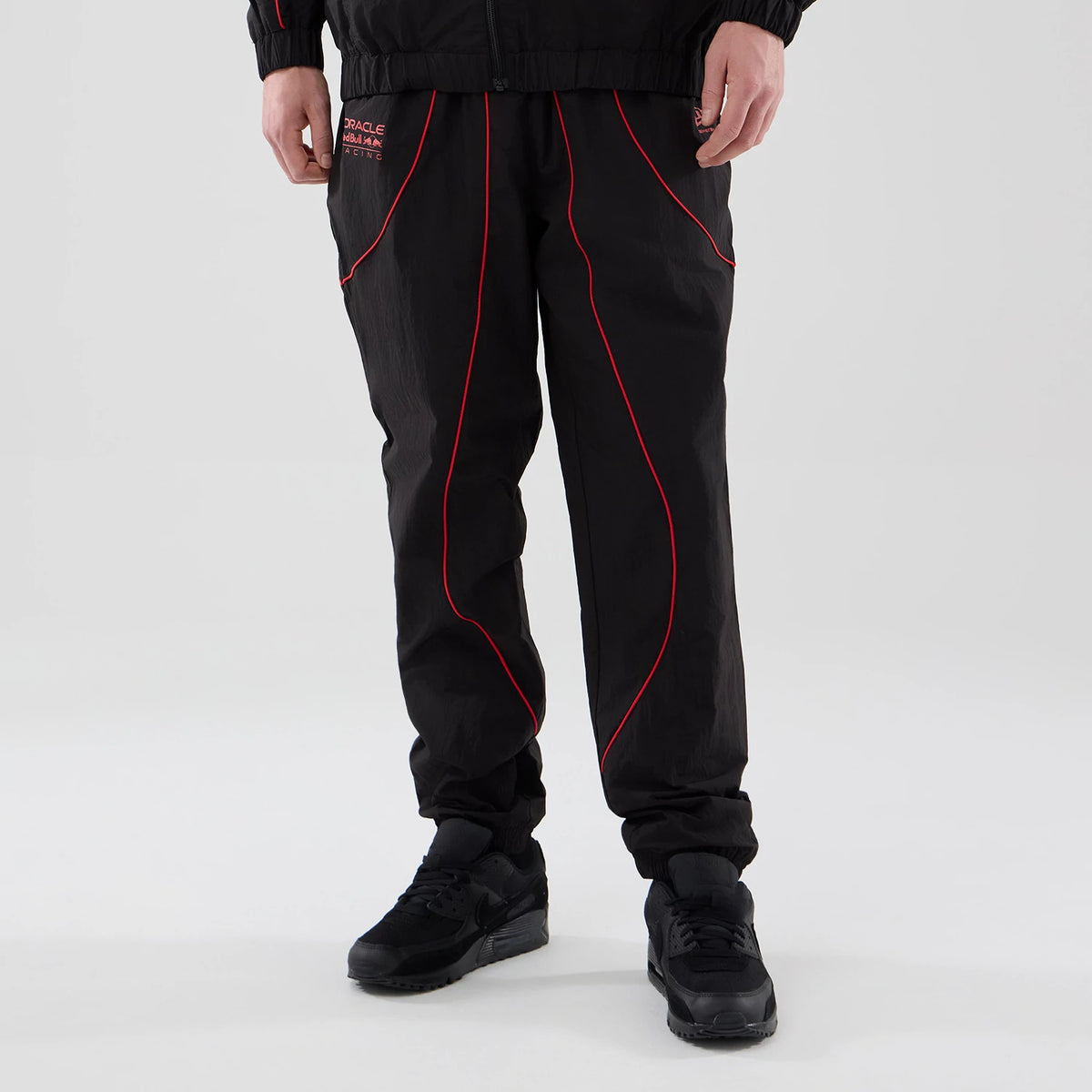 New Era Red Bull Racing Piping Track Black Track Pants, front view on a model showing red piping details curving around the front of the legs, Oracle Red Bull Racing lettering on the upper left thigh, and a relaxed fit down to the ankles.