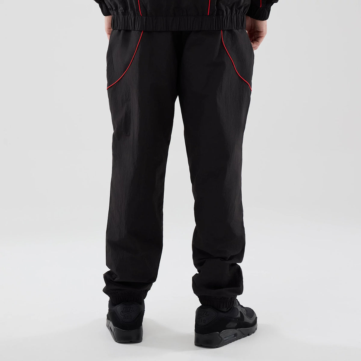 New Era Red Bull Racing Piping Track Black Track Pants, rear view on a model showing red piping details along the articulated knee design, elasticised cuffs at the ankles, and a relaxed fit throughout the black fabric.