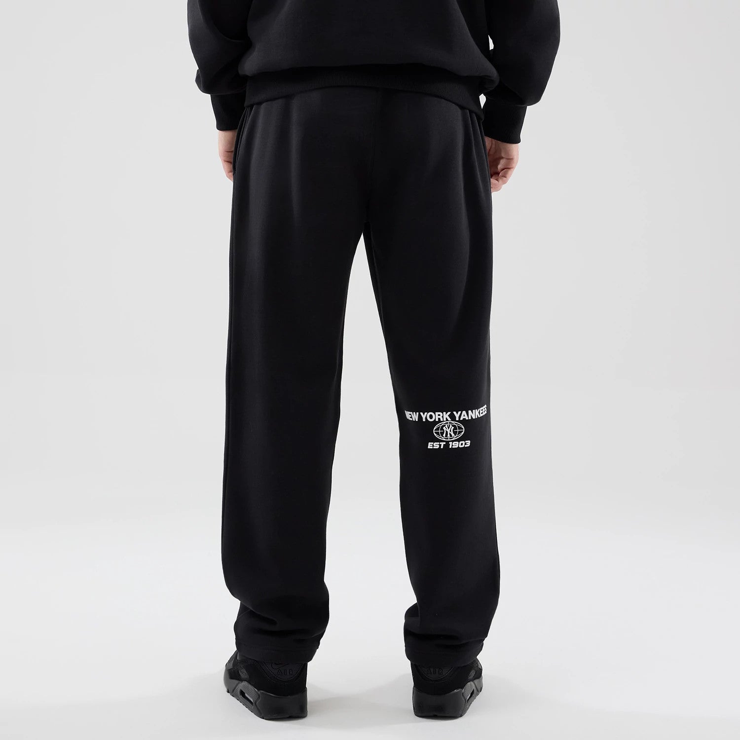 New Era New York Yankees MLB Logo Graphic Black Straight Leg Joggers, rear view on a model showing the white New York Yankees logo with 'Est 1903' under it on the left leg, and an elastic waistband with a relaxed fit.