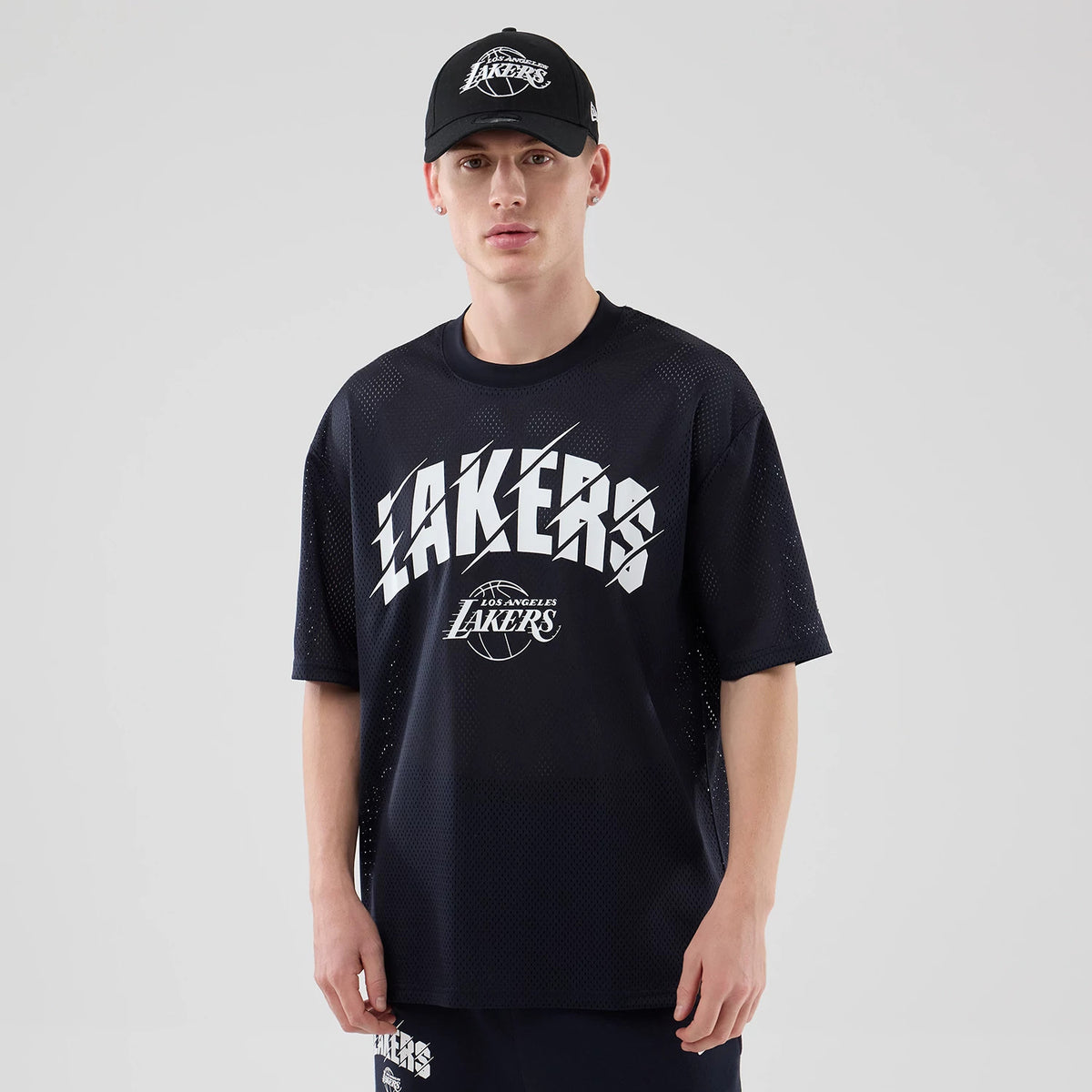 New Era LA Lakers NBA Core Plus Mesh Navy Oversized T-Shirt, front view on a model wearing a matching cap, featuring large white Lakers lettering across the chest with an embroidered NBA logo below.