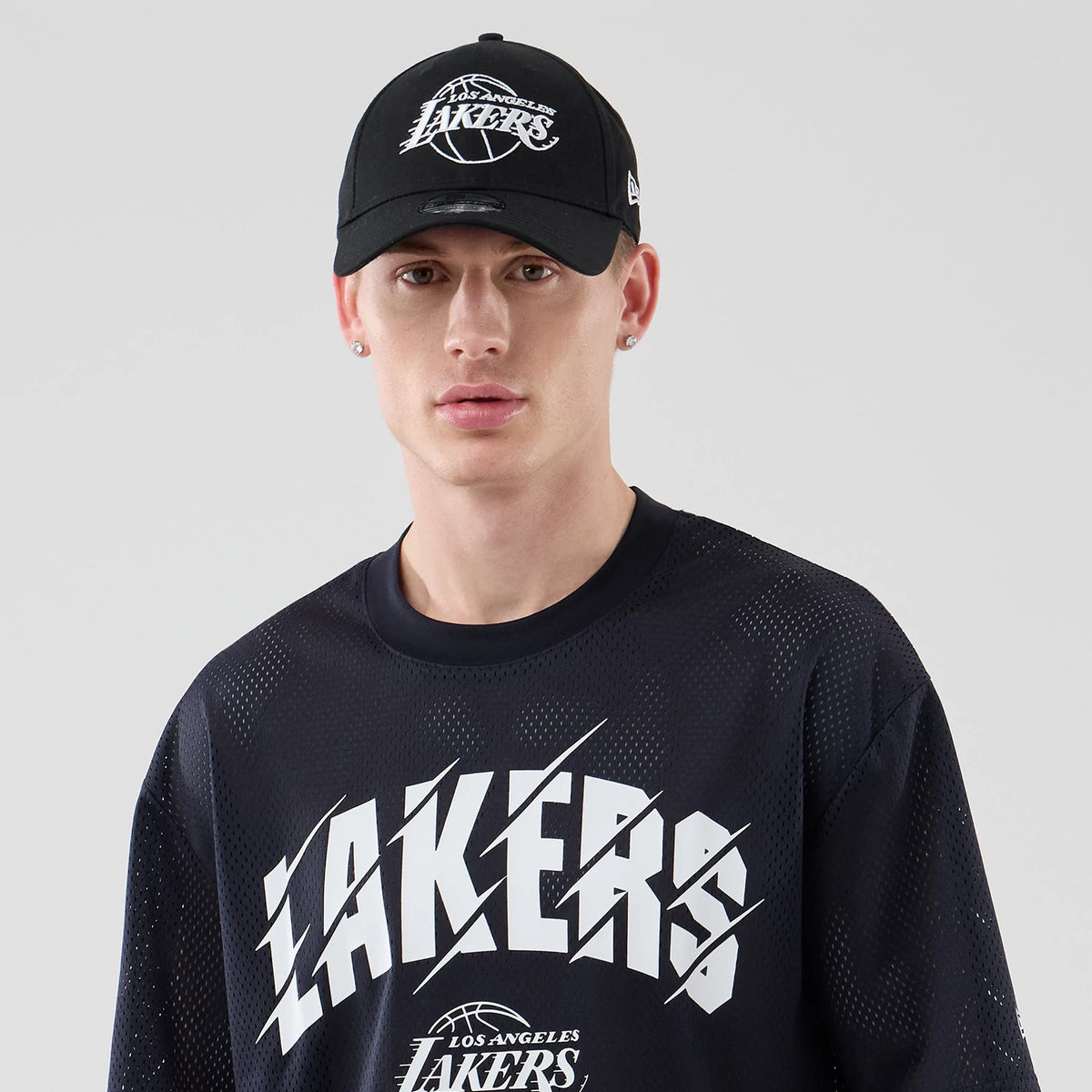 New Era LA Lakers NBA Core Plus Mesh Navy Oversized T-Shirt, front view on a model with a matching cap, showing white Lakers lettering across the chest, a smaller Los Angeles Lakers logo below, and detailed mesh fabric texture throughout.