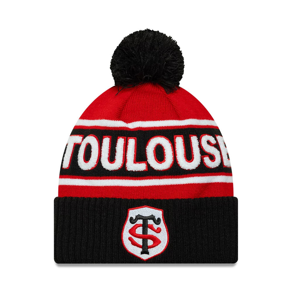 New Era Stade Toulousain Sport Red Cuff Knit Beanie Hat, front view showing white TOULOUS lettering across the red crown with black stripes, a large black pom-pom on top, and the embroidered Stade Toulousain logo centred on the black cuff.