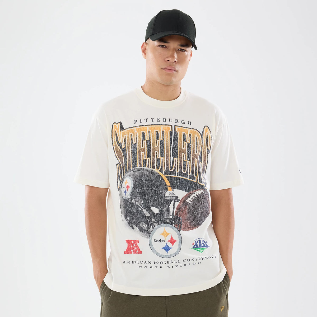 New Era Pittsburgh Steelers NFL Sport Classic Cream T-Shirt, front view on a model wearing a black cap, showcasing a large Steelers graphic in yellow and black with team logos, a football and helmet graphic across the chest.