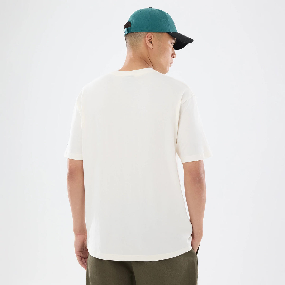 New Era Philadelphia Eagles NFL Sport Classic Cream T-Shirt, back view on a model wearing a teal cap, showing the plain cream fabric with short sleeves, a simple crew neckline and an untucked hemline, paired with olive green trousers.