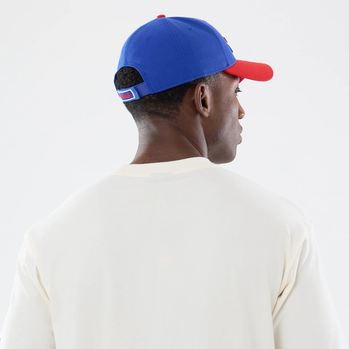 New Era Buffalo Bills NFL Sport Classic Cream T-Shirt, back view on a model wearing a blue cap with a red visor, showing the plain cream fabric with no graphics and a small embroidered Bills logo near the strap of the cap.