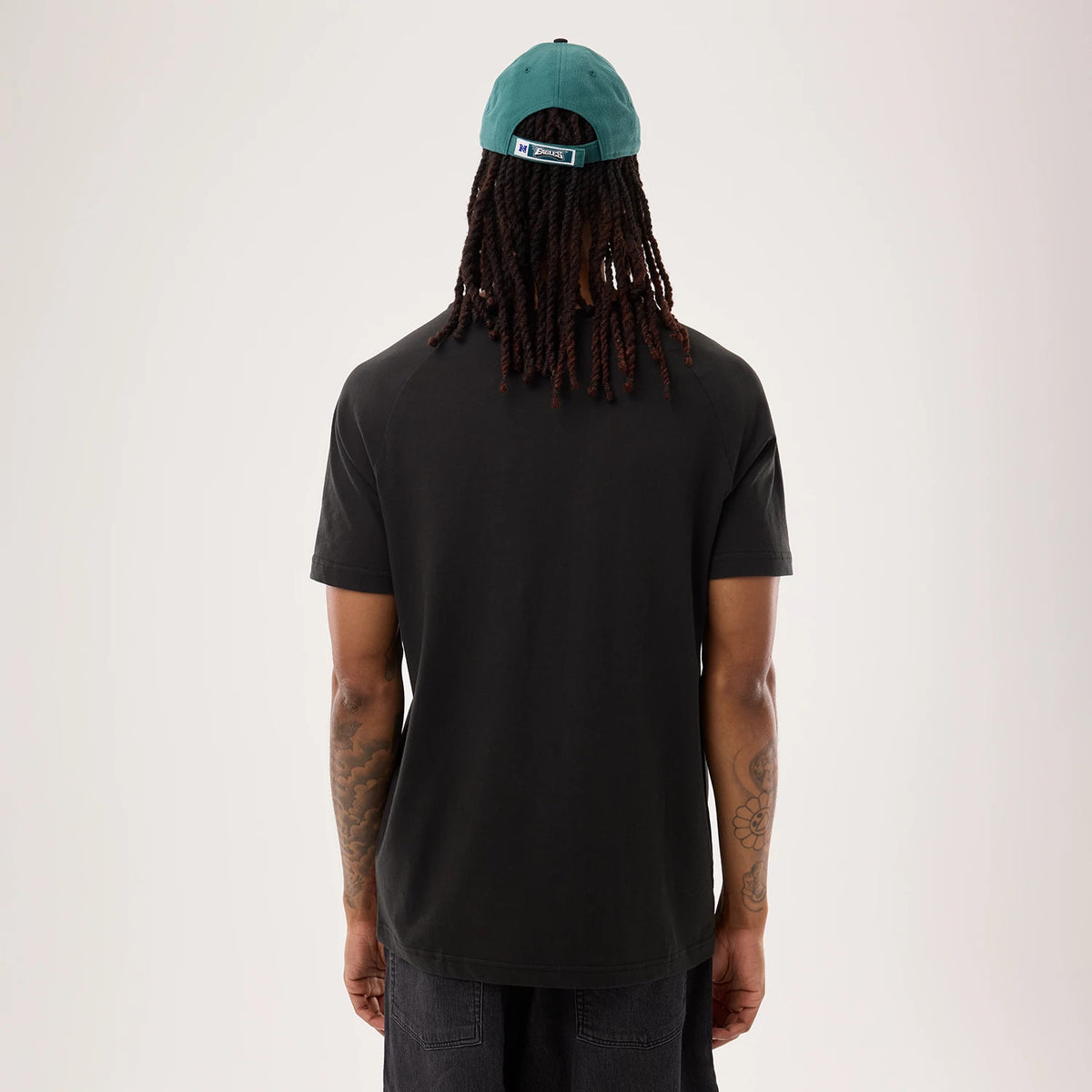 New Era Philadelphia Eagles NFL Sideline 2026 Black T-Shirt, back view on a model with a green cap showing the plain black back of the T-shirt, short sleeves, and a simple crew neckline. The cap features a white embroidered logo above the strap.