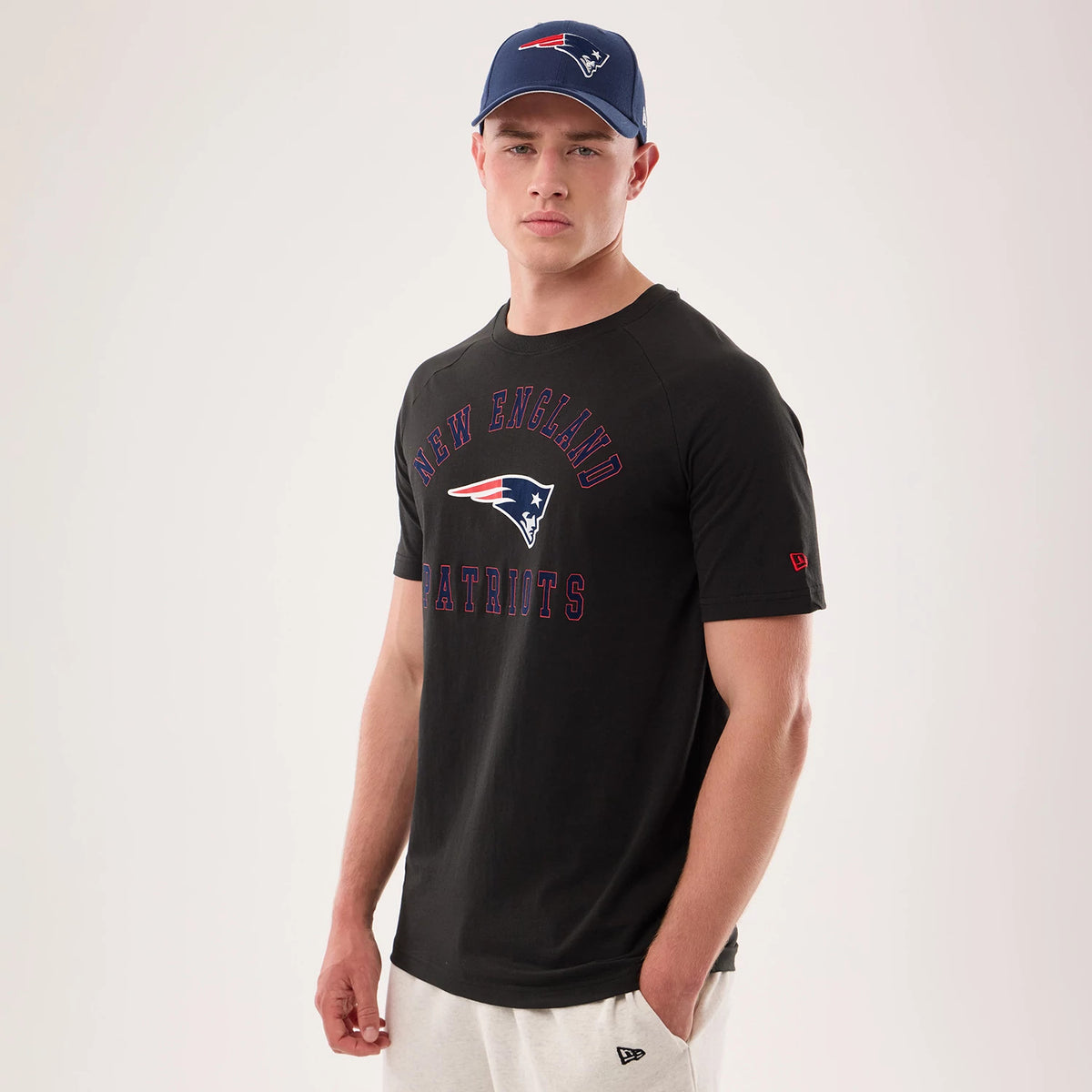 New Era New England Patriots NFL Sideline 2026 Black T-Shirt, front view on a model displaying New England lettering and team logo emblazoned across the chest, with a red embroidered New Era logo on the sleeve above light trousers.