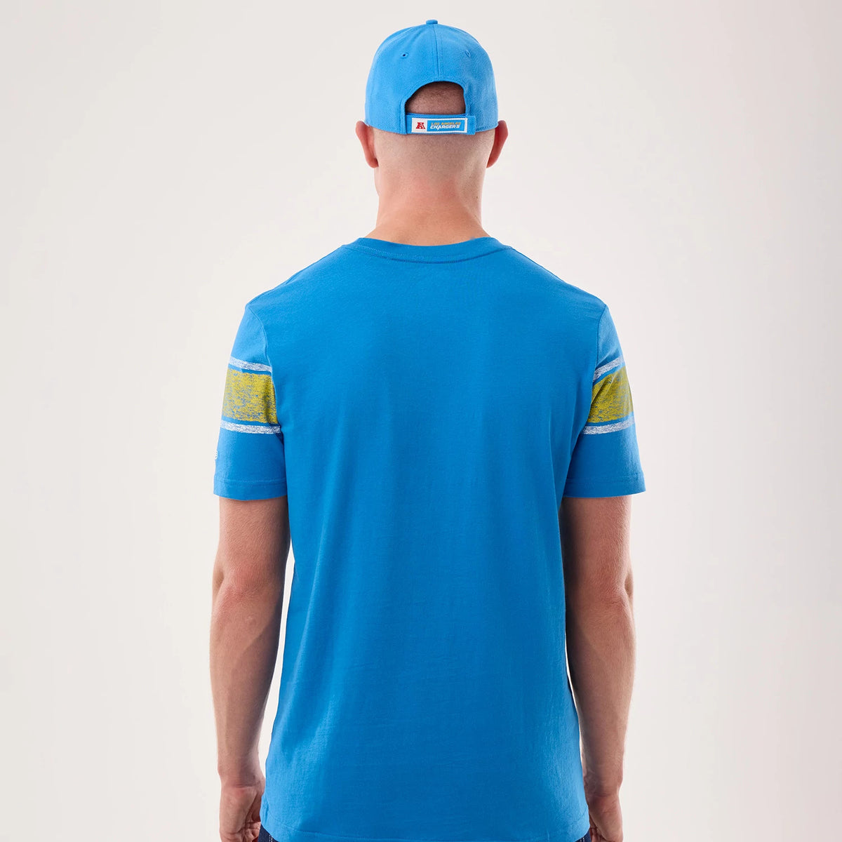 New Era LA Chargers NFL Sideline 2026 Blue T-Shirt, back view on a model with a blue cap, featuring yellow and white distressed stripes on the sleeves, a plain blue back, and the New Era logo embroidered on the cap's rear strap.