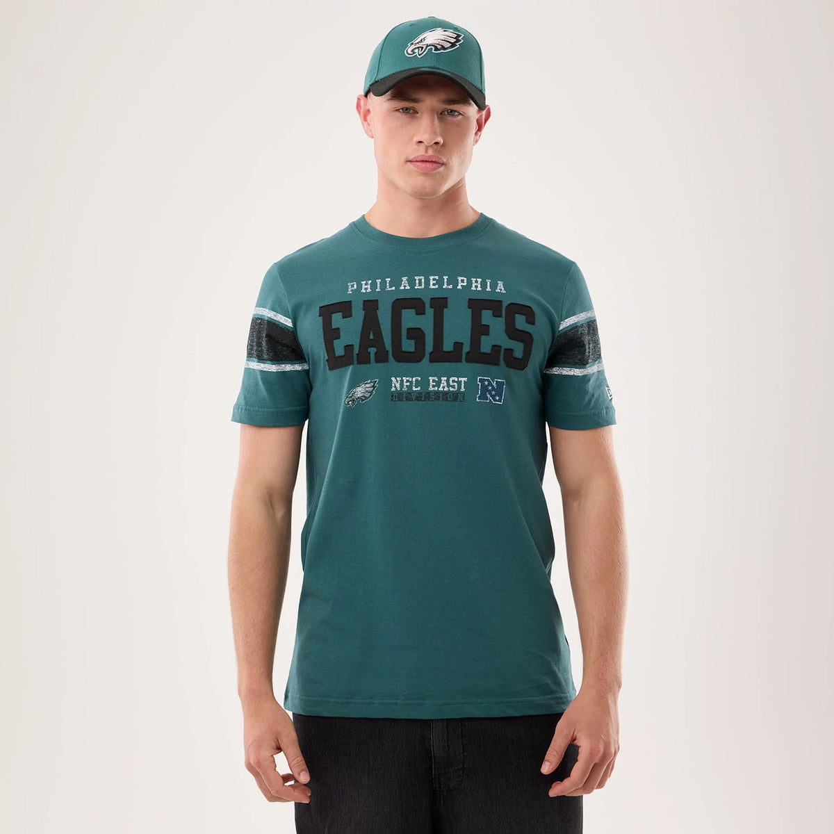 New Era Philadelphia Eagles NFL Sideline 2026 Dark Green T-Shirt, front view on a model with a matching green cap, showcasing large Eagles lettering on the chest above the NFC East text and team logo, with contrasting sleeve stripes.