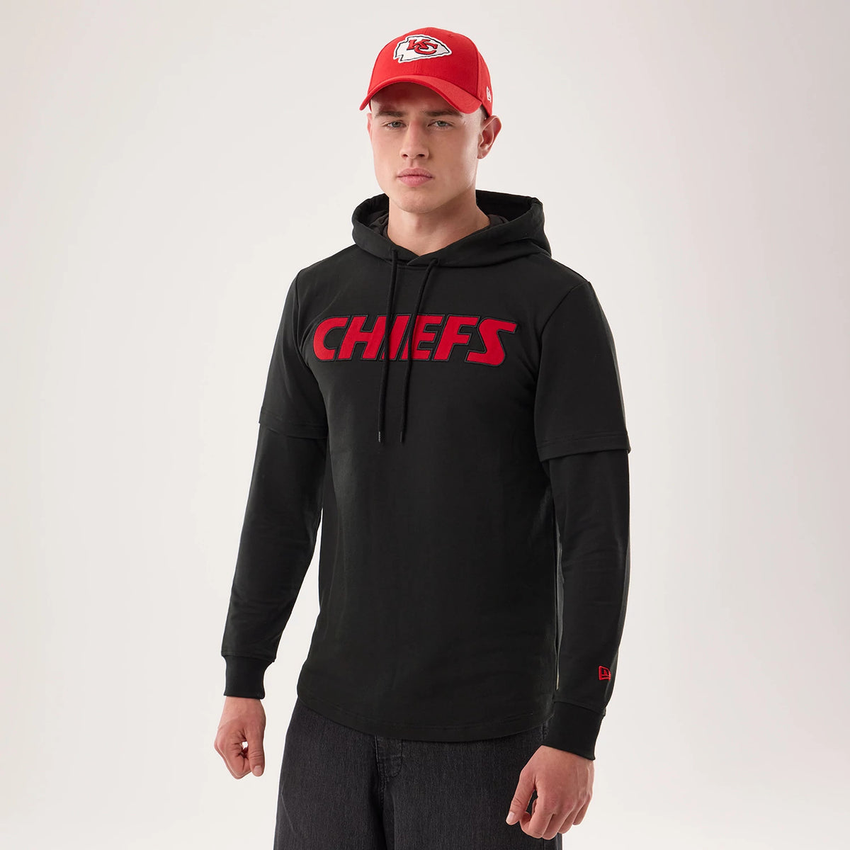 New Era Kansas City Chiefs NFL Sideline 2026 Black Double Sleeve Hoodie, front view on a model with a red cap, showing red CHIEFS lettering across the chest, double-layered sleeves, and a red embroidered New Era logo on the left cuff.