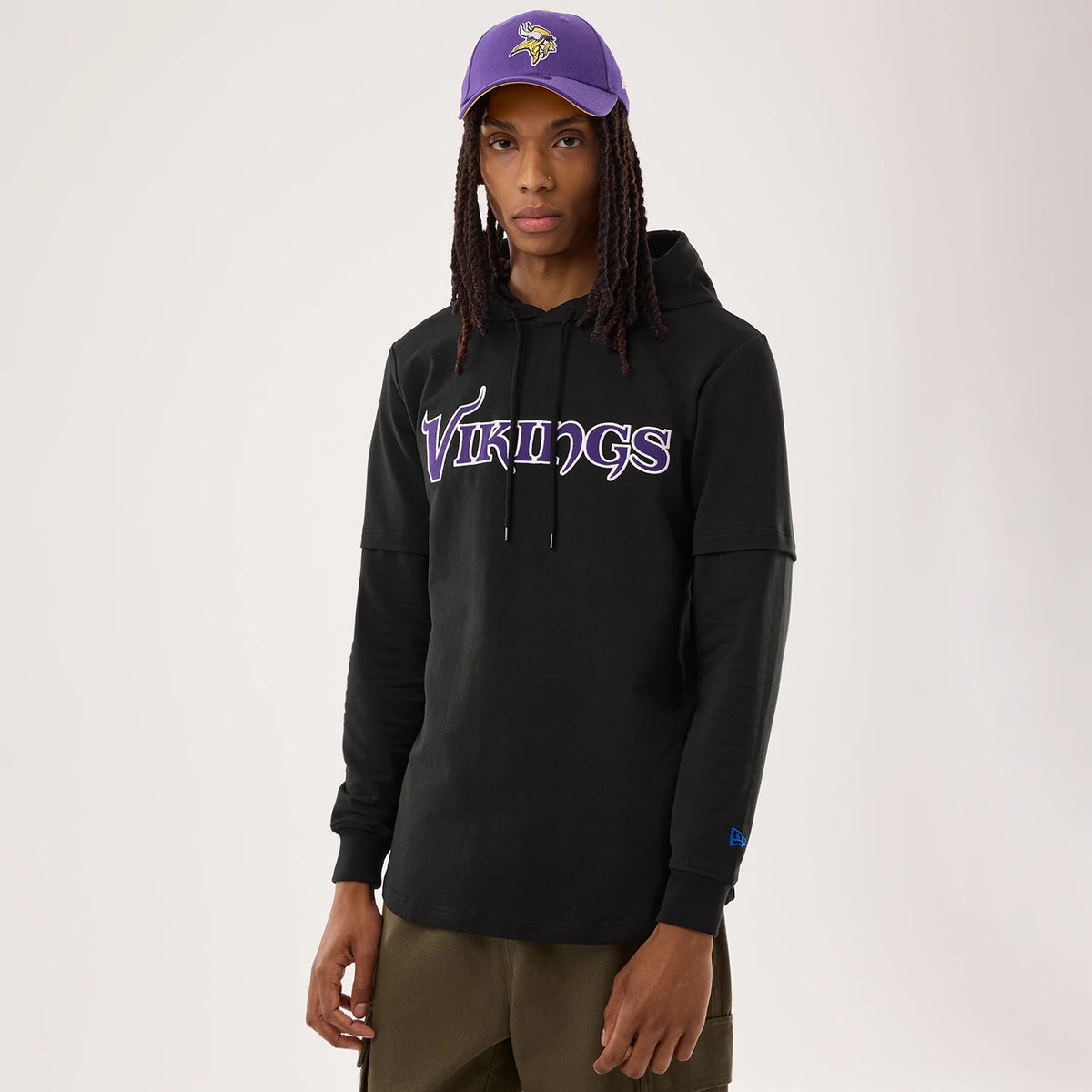 New Era Minnesota Vikings NFL Sideline 2026 Black Double Sleeve Hoodie, front view showing Vikings lettering across the chest in purple and white, worn by a model with olive trousers, featuring a small blue embroidered New Era logo on the cuff.