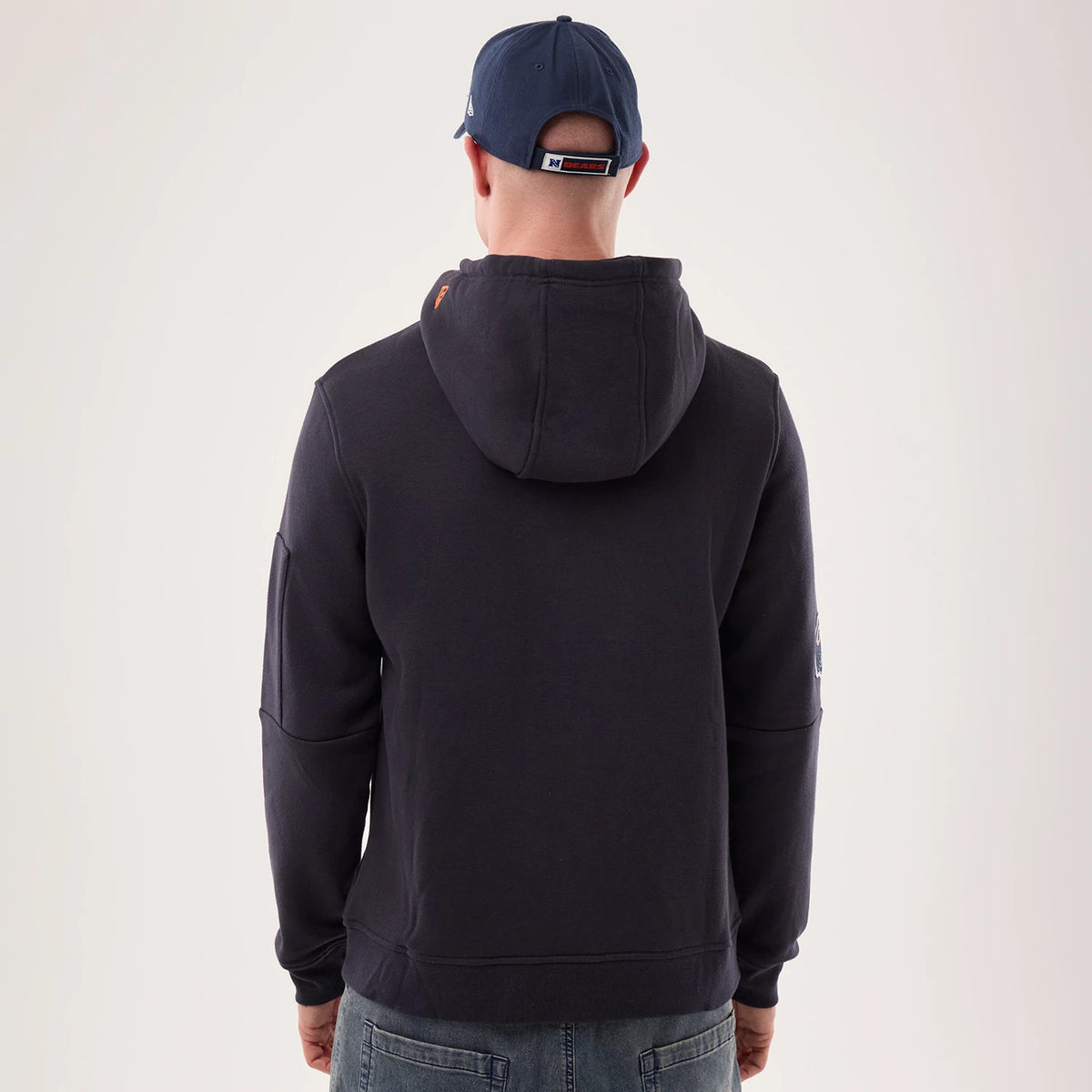 New Era Chicago Bears NFL Sideline 2026 Navy Pullover Hoodie, back view on a model with a navy cap, showing a plain navy back with a large hood, a orange New Era logo on the right shoulder and a small embroidered logo on the right sleeve.