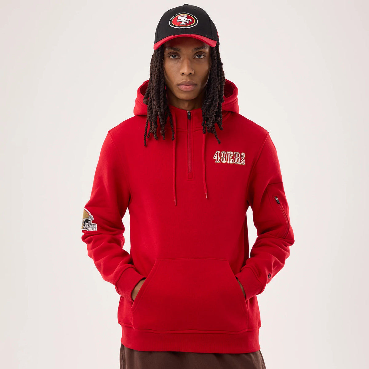 New Era San Francisco 49ers NFL Sideline 2026 Red Pullover Hoodie, front view on a model with a matching cap, featuring a red pullover design with embroidered 49ERS lettering on the chest, a team patch on the sleeve, and a hood with drawstrings.