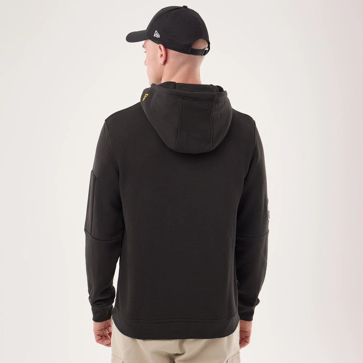 New Era Pittsburgh Steelers NFL Sideline 2026 Black Pullover Hoodie, back view on a model with beige trousers and a black cap, showing a simple black hood, a small yellow embroidered logo at the neck, and a seam detail on the sleeve.