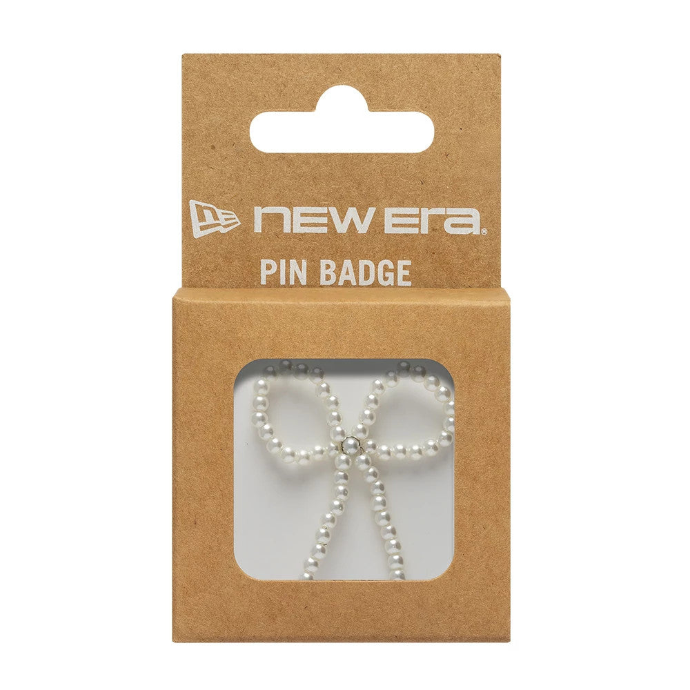 New Era Faux Pearl Bow White Pin Badge, front view showing a delicate bow shape made from small, faux pearls, with a single clear gemstone at the centre, packaged in a brown card holder featuring the New Era logo in white above.