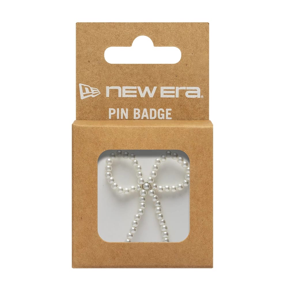 New Era Faux Pearl Bow White Pin Badge, front view showing a delicate bow shape made from small, faux pearls, with a single clear gemstone at the centre, packaged in a brown card holder featuring the New Era logo in white above.