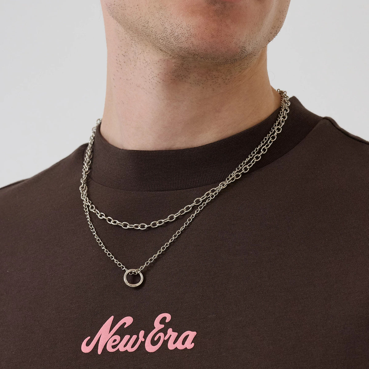 New Era Love Graphic Dark Brown T-Shirt, front view on-model showing the soft pink embroidered New Era logo prominently centred near the chest, complemented by layered silver chain necklaces against the dark brown fabric.