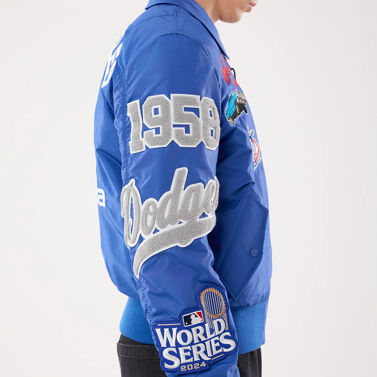 New Era LA Dodgers MLB X Alpha Blue Jacket, side view showing large grey embroidered 1958 and Dodgers lettering on the sleeve, colourful patches on the chest, and the MLB World Series 2024 embroidered logo near the cuff on the bright blue fabric.