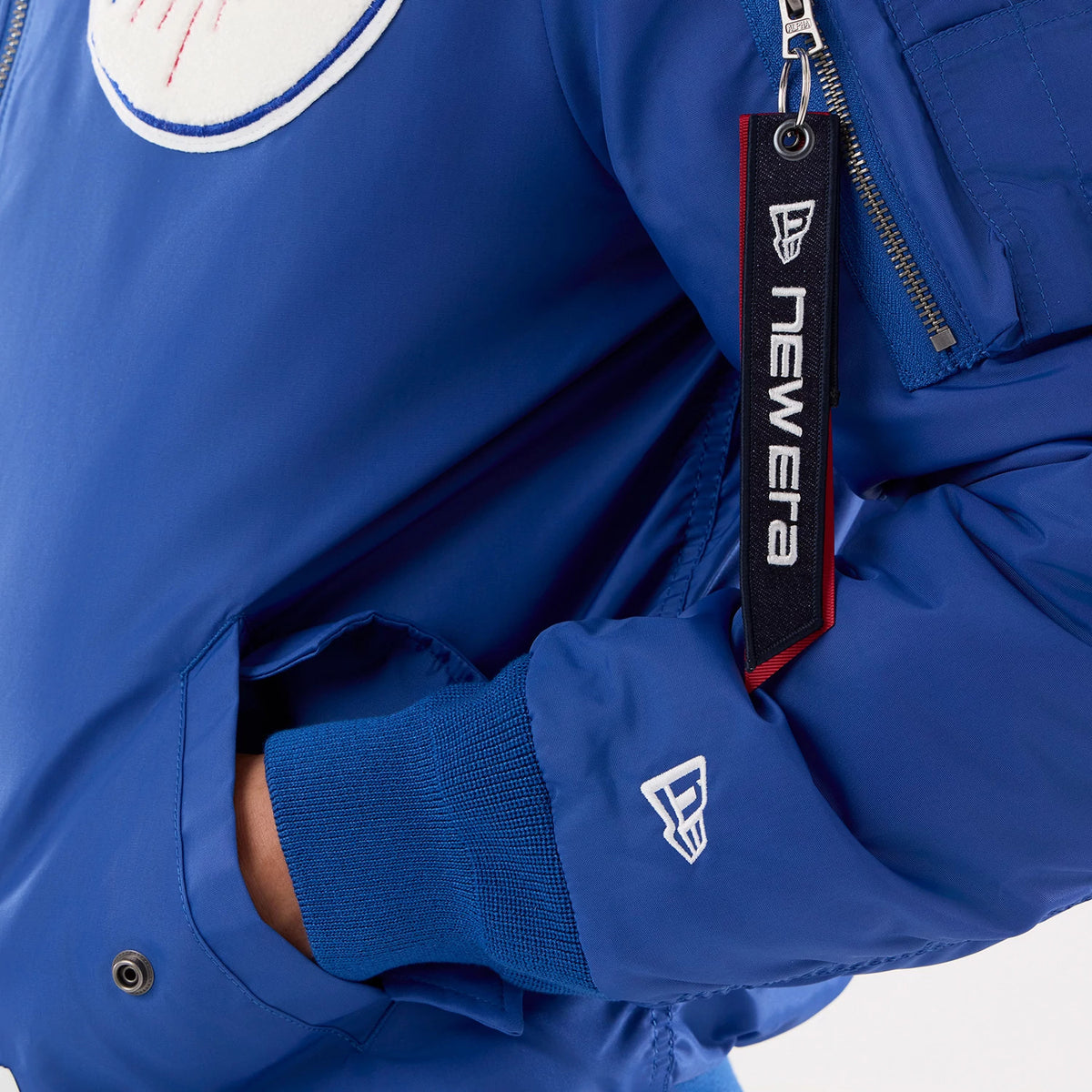 New Era LA Dodgers MLB X Alpha Blue Jacket, close-up view showing a blue sleeve with ribbed cuffs, an embroidered New Era logo on the wrist, an angled zipped pocket on the upper arm, and a black and red New Era tag attached to the zip pull.