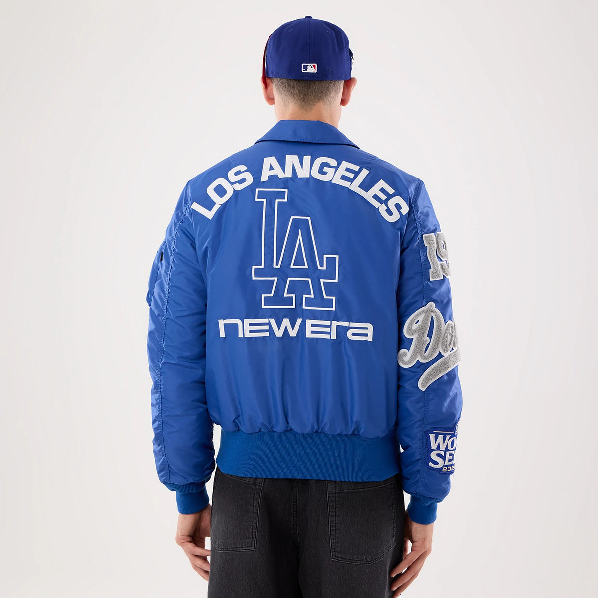 New Era LA Dodgers MLB X Alpha Blue Jacket, back view on a model with a matching cap showing embroidered Los Angeles lettering and large LA logo in white, New Era logo below, and detailed patches on the sleeves, including Dodgers script.