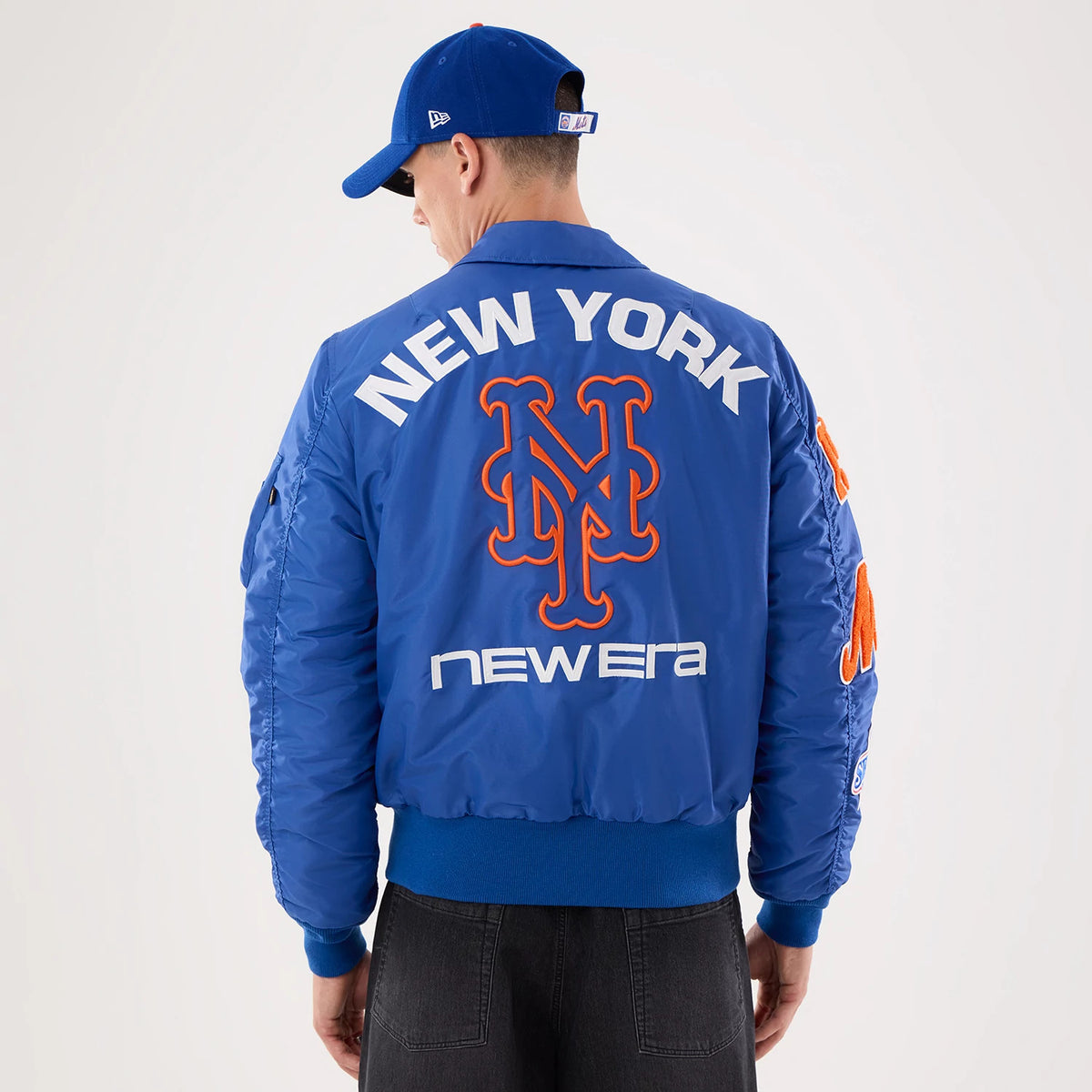 New Era New York Mets MLB X Alpha Blue Jacket, back view on a model with a blue cap, featuring large white New York lettering and an orange-accented Mets logo, the New Era logo at the bottom, and additional orange details on the sleeves.