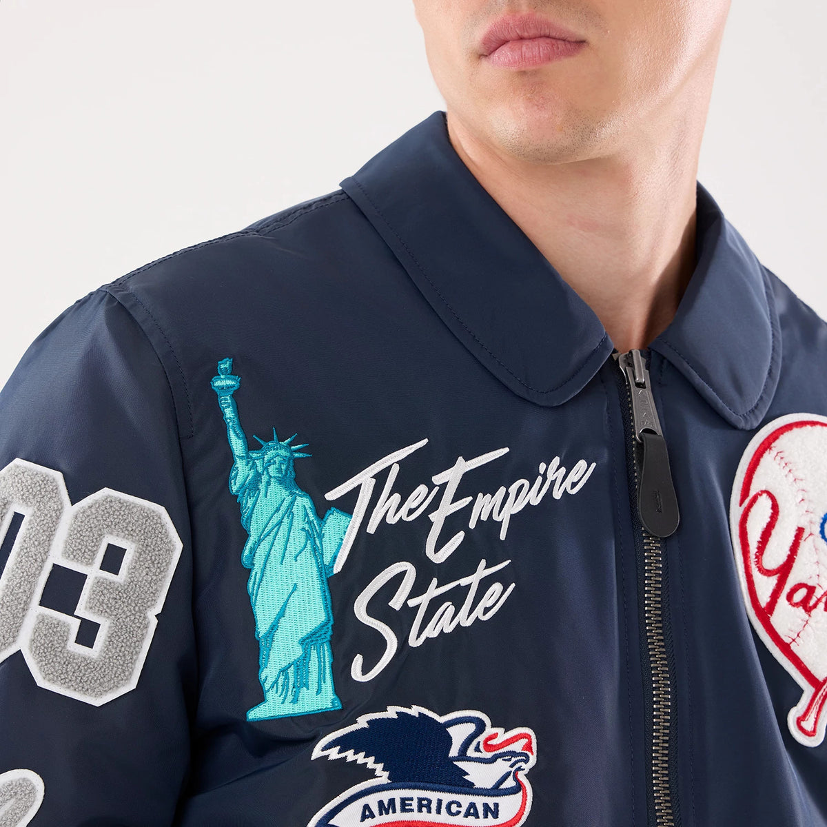 New Era New York Yankees MLB X Alpha Navy Jacket, front view showing multiple embroidered patches including the Statue of Liberty in turquoise, The Empire State in white lettering, and a large baseball-themed patch, with a zip closure.