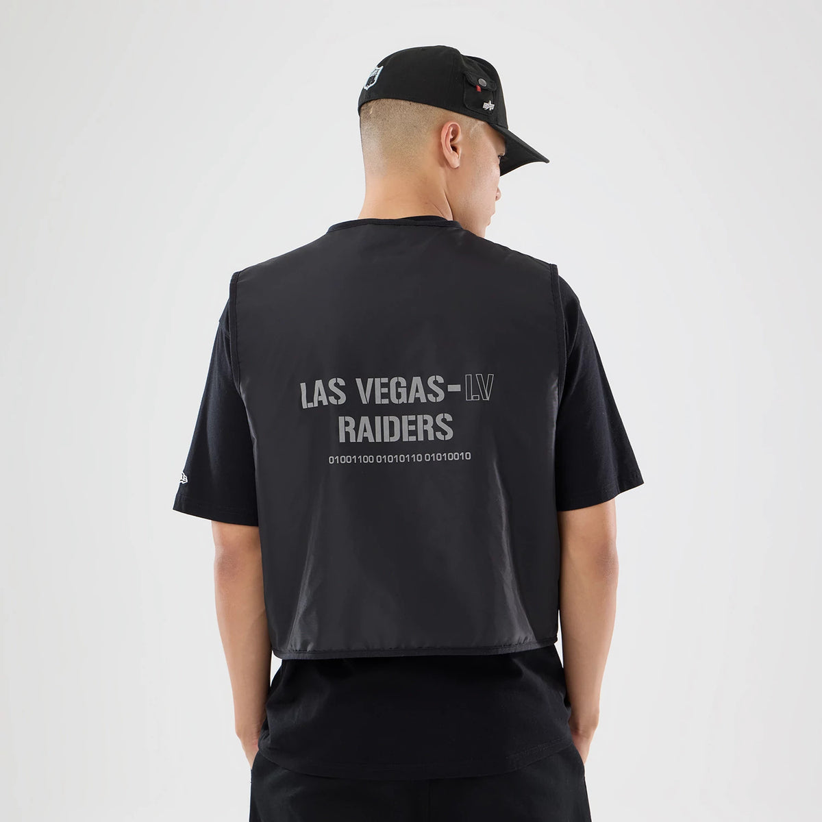 New Era Las Vegas Raiders NFL X Alpha Black Bomber Jacket & Tactical Vest, back view on a model showing the large grey printed Las Vegas-LV Raiders lettering with binary code beneath, worn over a black T-shirt and paired with a black cap.