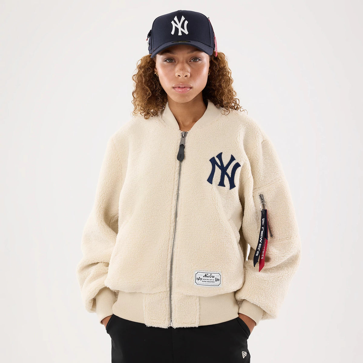 New Era New York Yankees MLB X Alpha Cream Borg Jacket, front view on a model showing the navy embroidered NY Yankees logo on the upper left chest, a central zip closure, and a New Era patch on the lower right hem, paired with a matching cap.