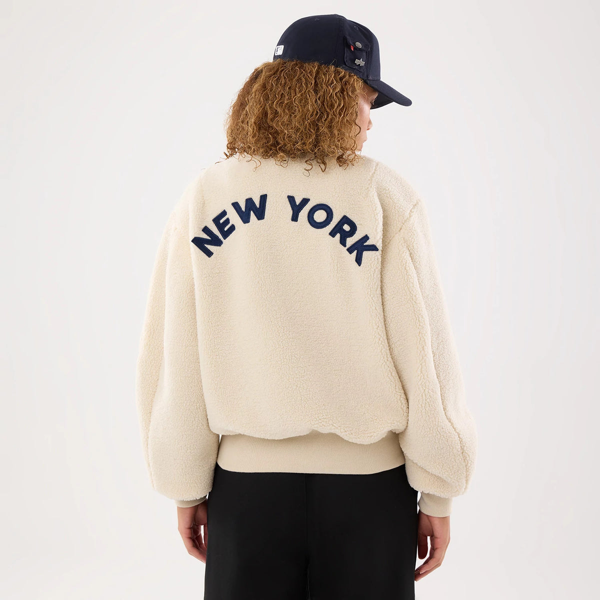 New Era New York Yankees MLB X Alpha Cream Borg Jacket, back view on a model wearing a navy cap, showcasing large New York lettering in raised navy blue across the upper back of the textured cream jacket with ribbed cuffs and hem.