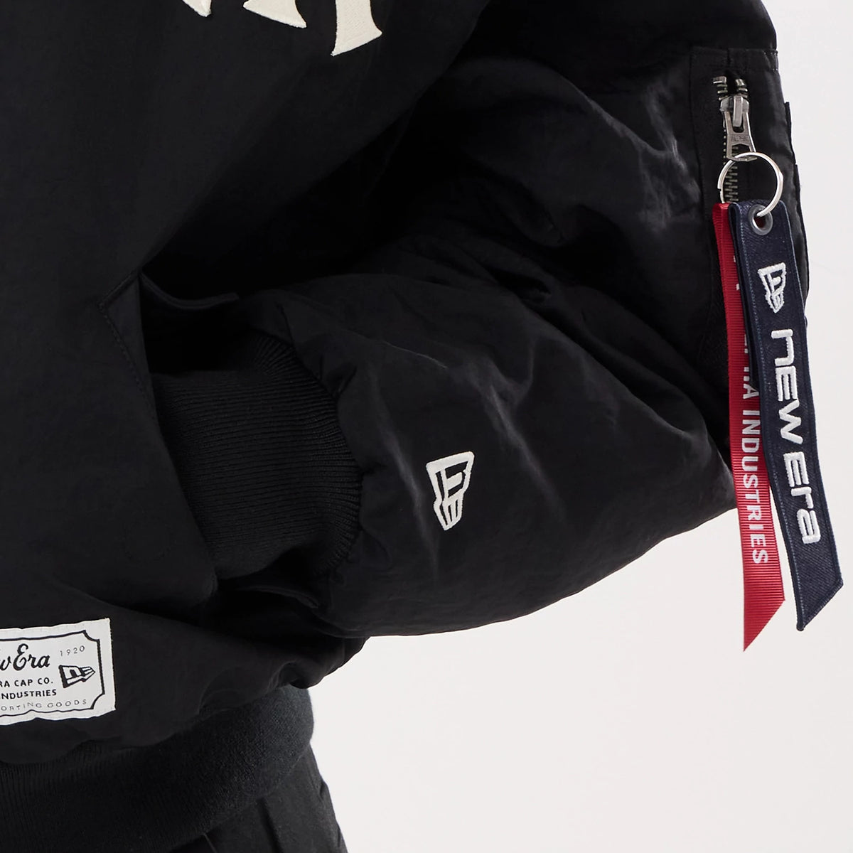 New Era New York Yankees MLB X Alpha Black Jacket, side view highlighting a white embroidered New Era logo on the sleeve, a black ribbed cuff and a sleeve pocket with a zip closure, adorned with navy and red branded tags hanging from the zip.