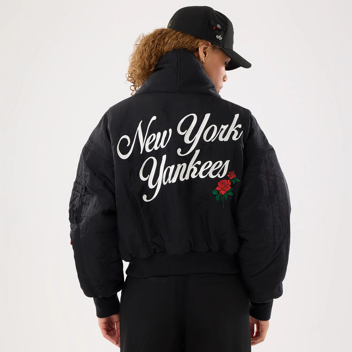 New Era New York Yankees MLB X Alpha Black Jacket, back view on a model with a black cap, featuring white embroidered New York Yankees lettering across the back and a detailed red rose graphic below the text on the black fabric.