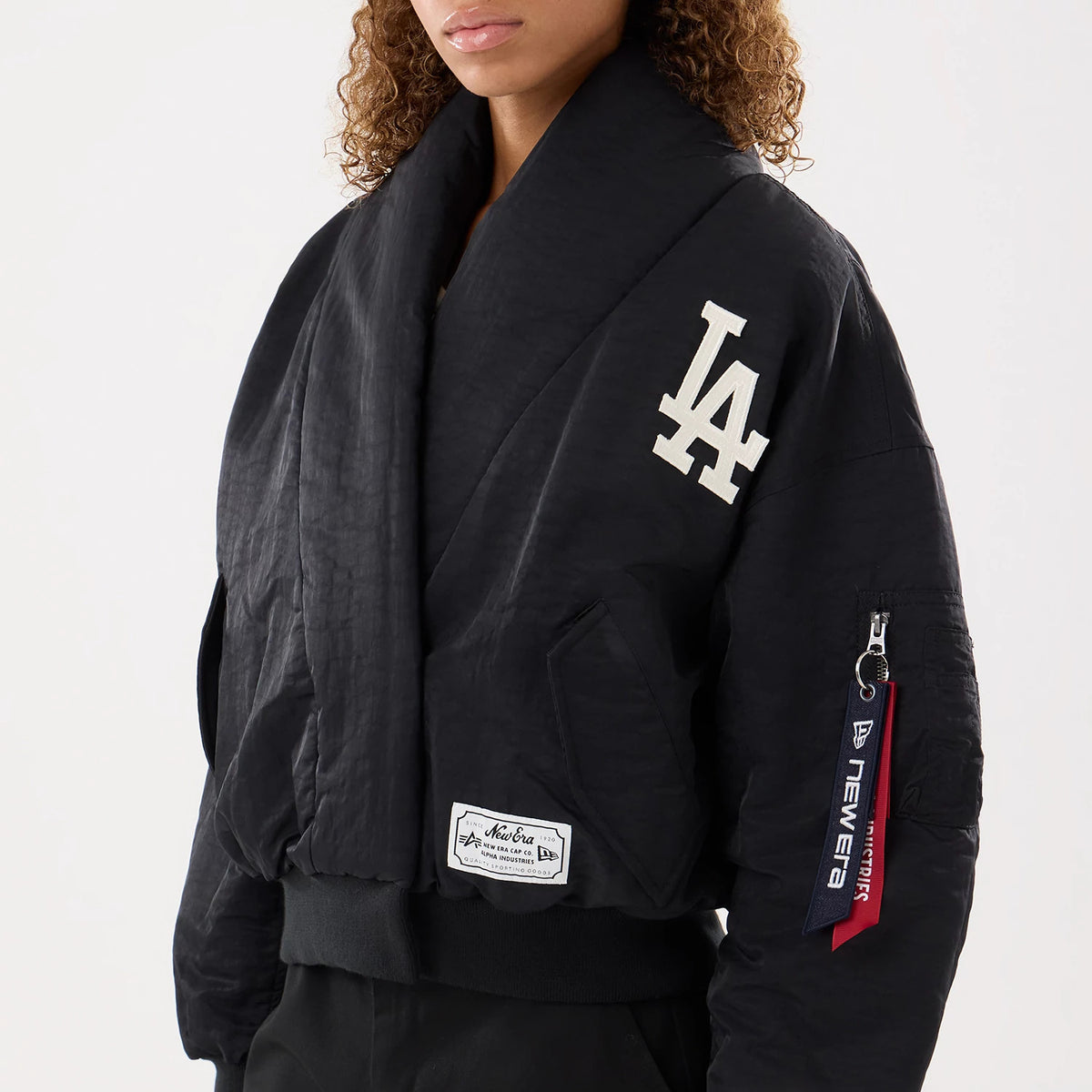 New Era LA Dodgers MLB X Alpha Black Jacket, front view on-model showing a prominent white embroidered LA logo on the left chest, a ribbed black hem, a New Era patch near the bottom, and a zip pocket with branded tags on the sleeve.