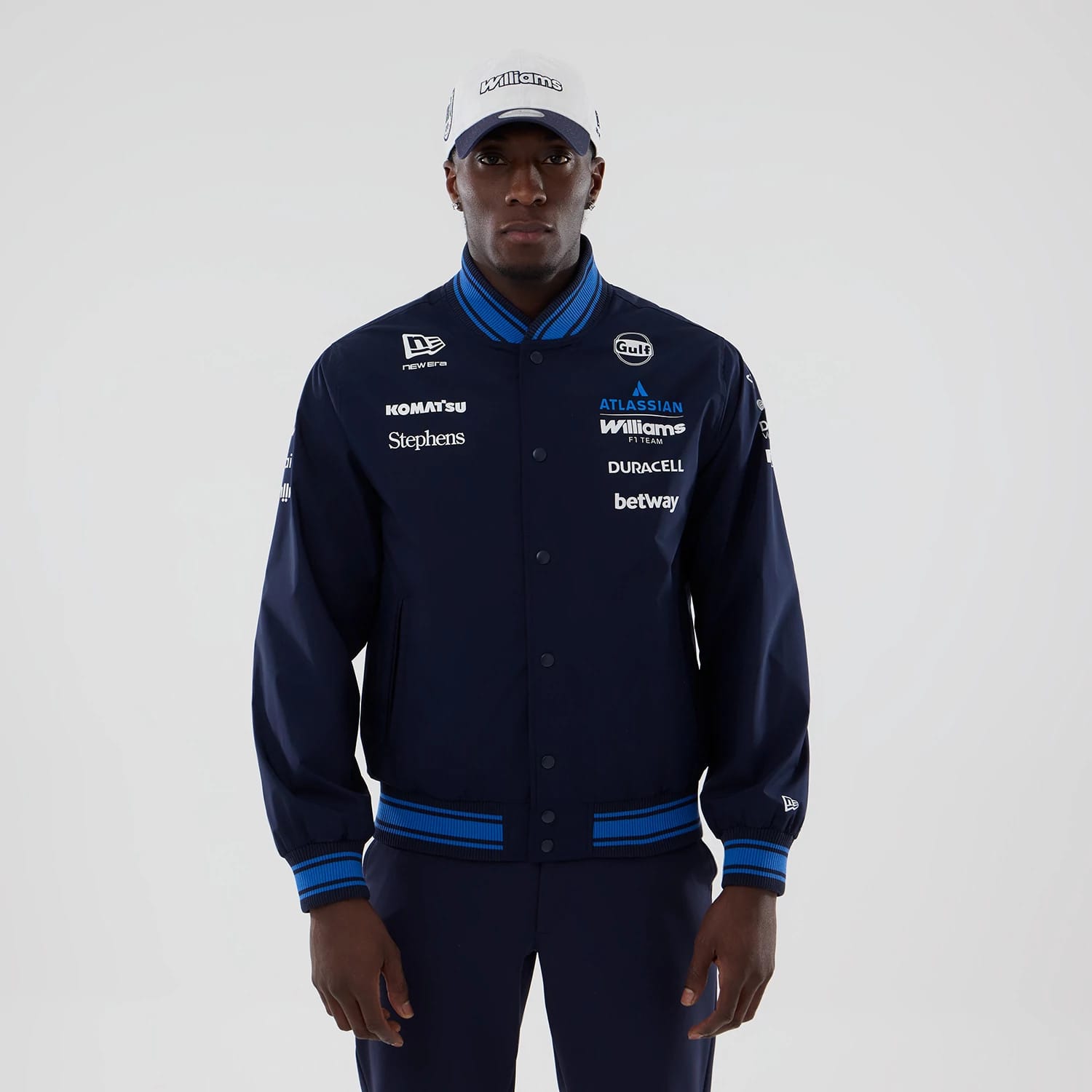 New Era Williams Navy Varsity Jacket, front view on-model showcasing a navy jacket with multiple white and blue sponsor logos embroidered on the chest and sleeves, a striped ribbed collar, hem, and cuffs.