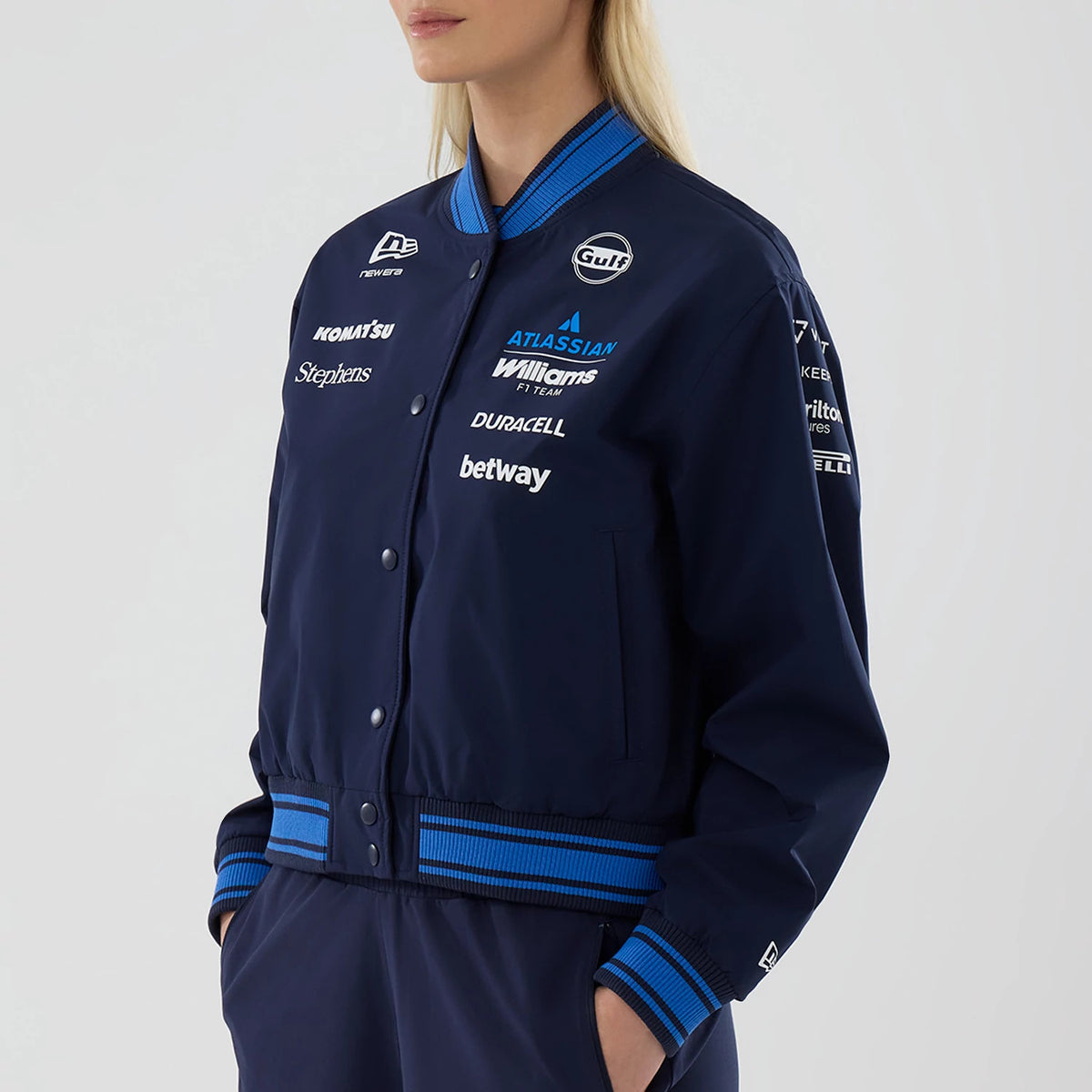 New Era Womens Williams Navy Varsity Jacket, front view on-model showcasing a navy jacket with multiple embroidered sponsor logos on the chest and sleeves, striped blue and navy ribbed collar, cuffs, and hem, and button closure down the front.