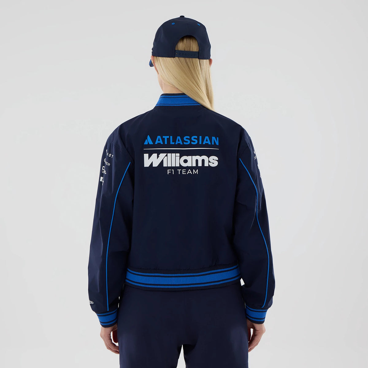 New Era Womens Williams Navy Varsity Jacket, back view on a model with a matching cap, showing large white Williams F1 Team lettering across the upper back, Atlassian logo above it, and blue stripes down the sleeves and around the cuffs.