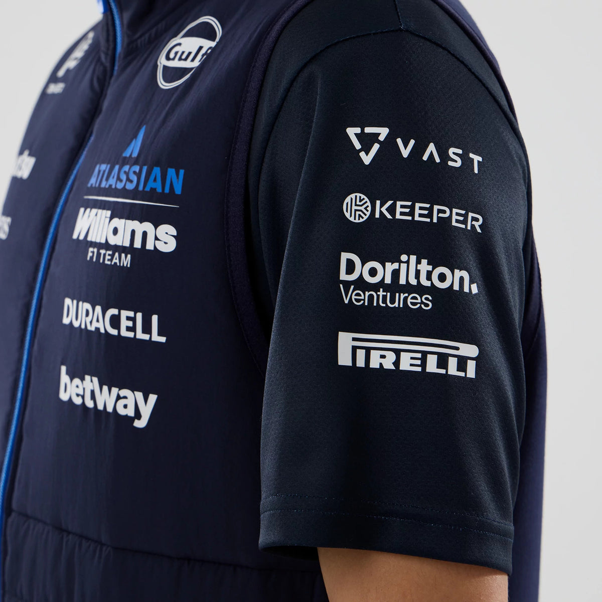 New Era Williams Navy Gilet, side view on a model with prominent sponsor logos including Gulf, Atlassian, and Williams F1 Team on the chest, complemented by a blue zipper and sleeveless design in dark navy fabric.