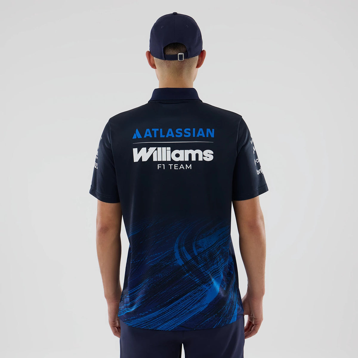 New Era Williams Navy Tech Polo T-shirt, back view on a model with a navy cap, showcasing white embroidered Williams F1 team lettering across the upper back and an Atlassian logo above, complemented by dynamic blue streaks at the lower back.