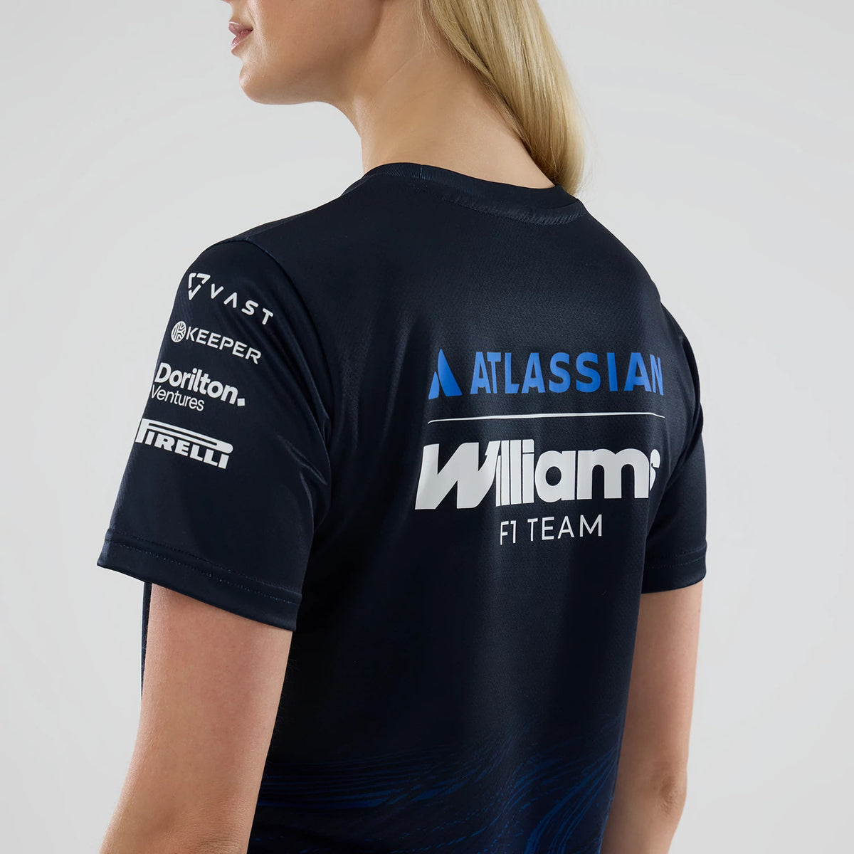 New Era Williams Womens Navy Tech T-shirt, back view on a model displaying large Williams F1 Team lettering predominantly across the upper back, with additional sponsor logos like Atlassian, VAST, and Pirelli arranged vertically on the sleeves.