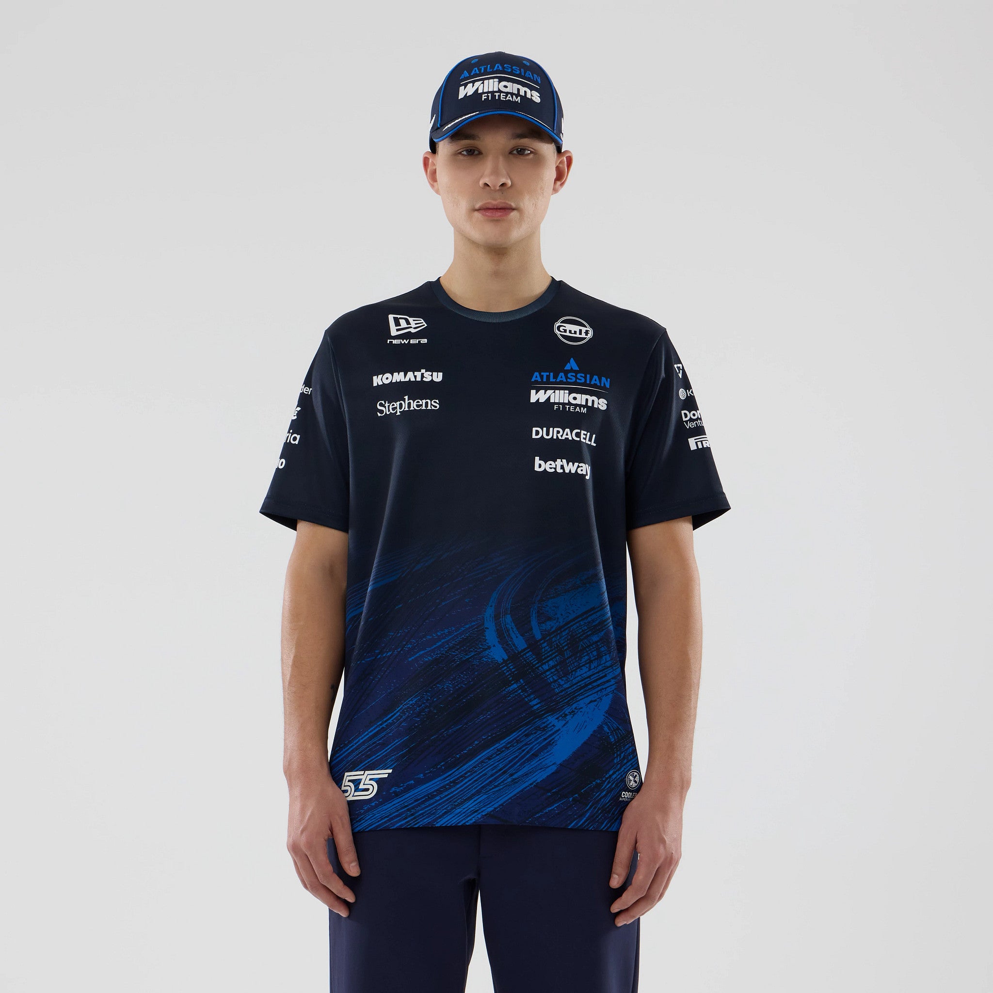 New Era Williams Carlos Sainz Navy Tech T-shirt, front view on a model paired with a blue cap, displaying multiple sponsor logos printed across the chest and sleeves, with a dynamic blue graphic pattern at the bottom.