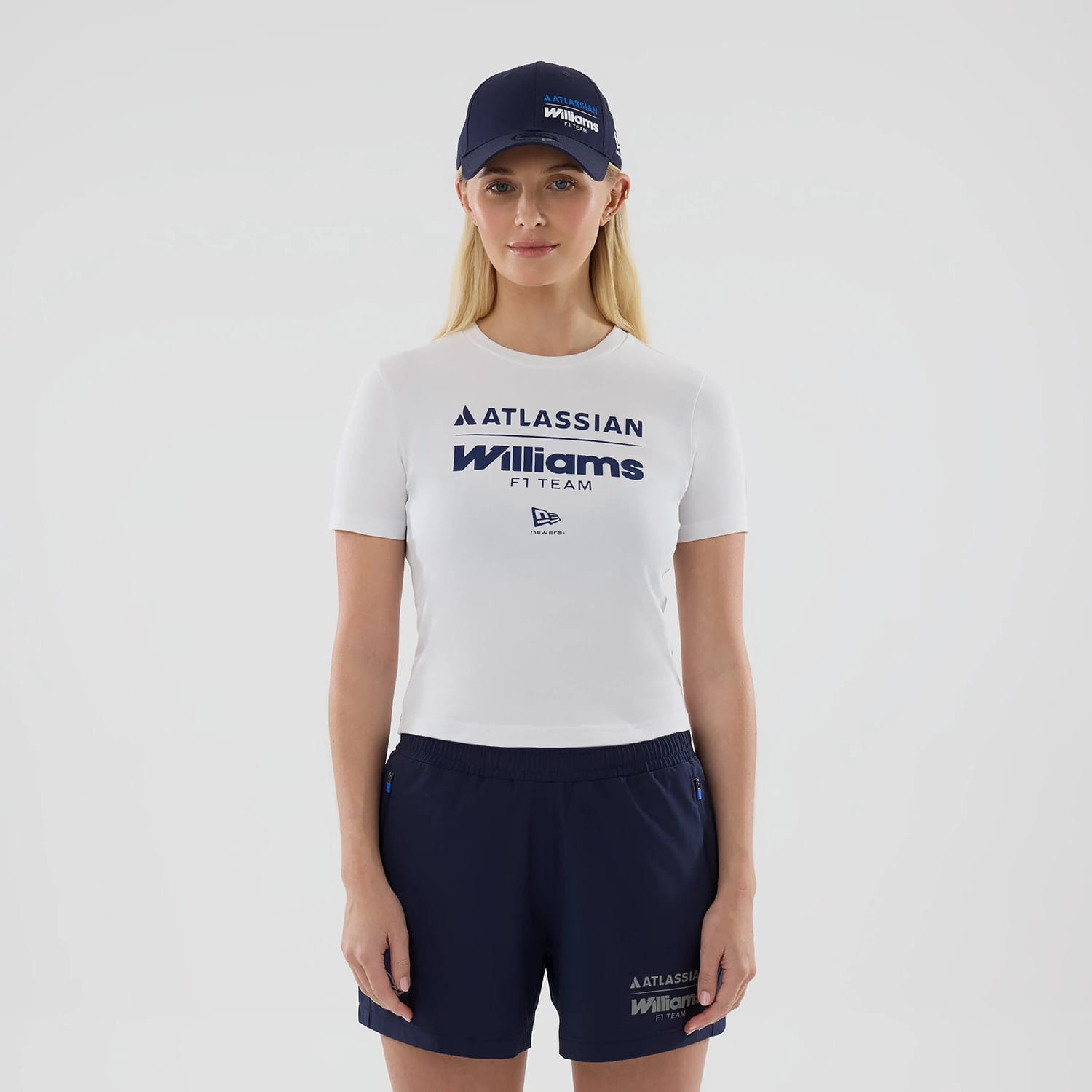 New Era Williams Womens White T-Shirt, front view on a model paired with a navy cap and navy shorts, featuring embossed Williams F1 Team lettering beneath an Atlassian logo, and an embroidered New Era flag logo centred below.