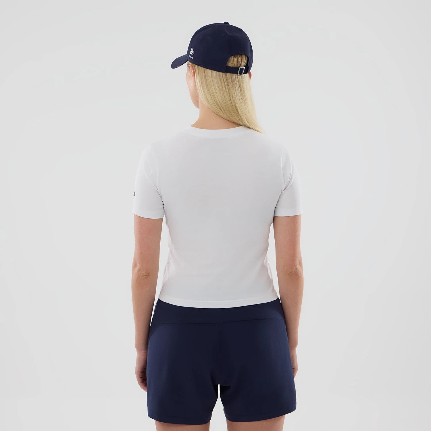 New Era Williams Womens White T-Shirt, back view on a model wearing a dark cap, showing the plain white short-sleeve design with a simple round neckline and a straight hem, complemented by navy shorts for a casual look.