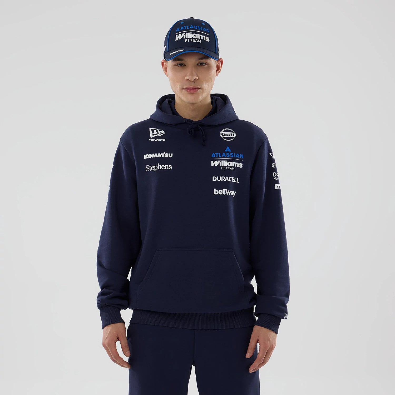 New Era Williams Replica Navy Pullover Hoodie, front view on a model styled with a matching cap, featuring multiple sponsor logos including embroidered New Era and Gulf on the chest, with a large front pocket and ribbed cuffs and hem.