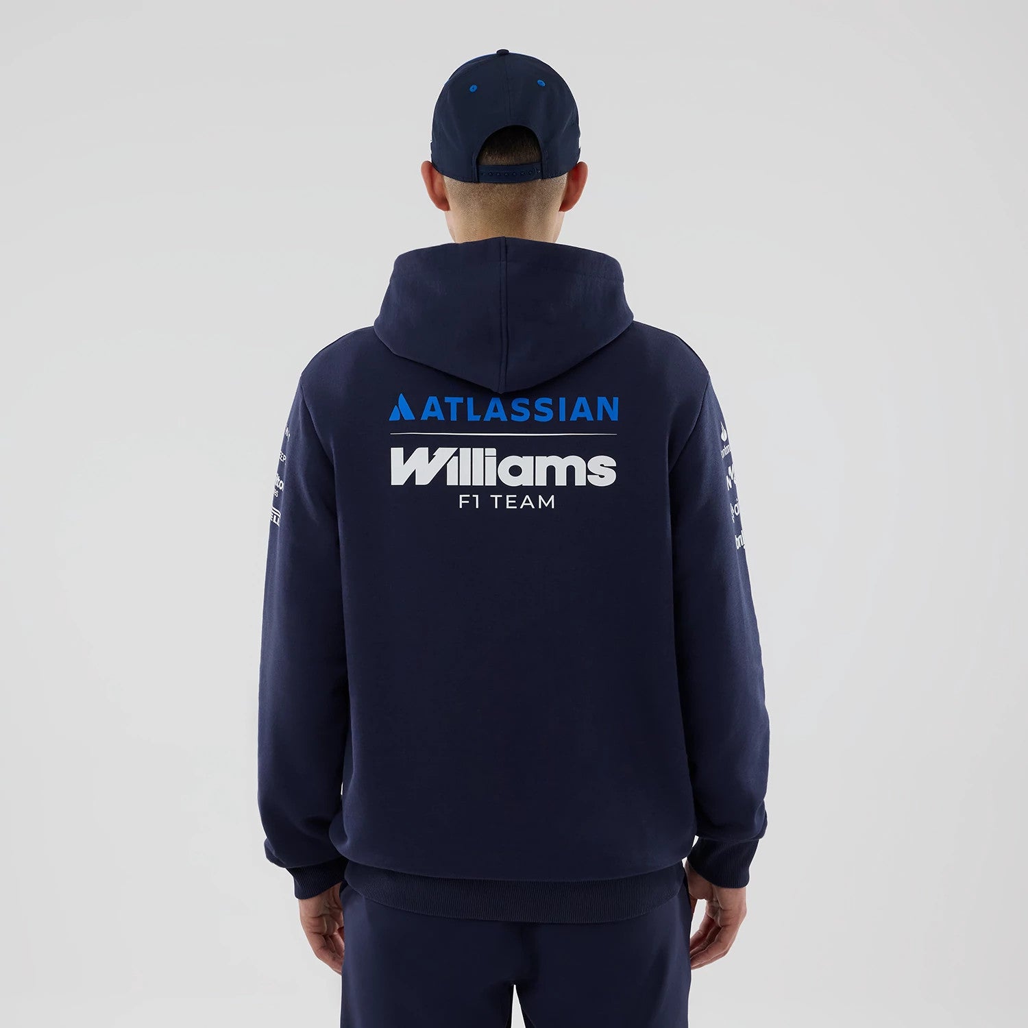 New Era Williams Replica Navy Pullover Hoodie, back view on a model with a matching navy cap, showing a large blue Atlassian logo above the white Williams F1 Team text across the back and branding along the sleeves.