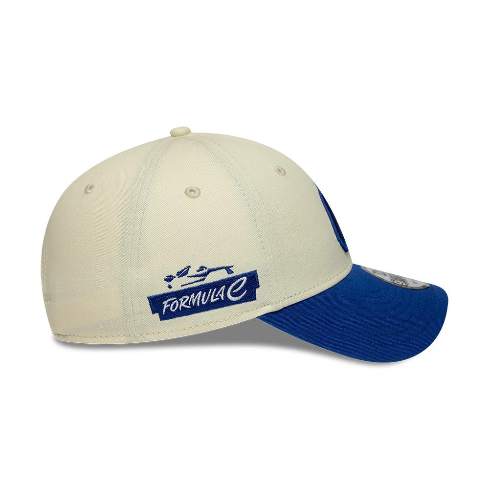 FIA Formula E Cream New Era 9FORTY Adjustable Cap, side view showing the blue embroidered Formula E logo on the cream crown, curved blue visor, and a 9FORTY size sticker partially on the top of the visor.