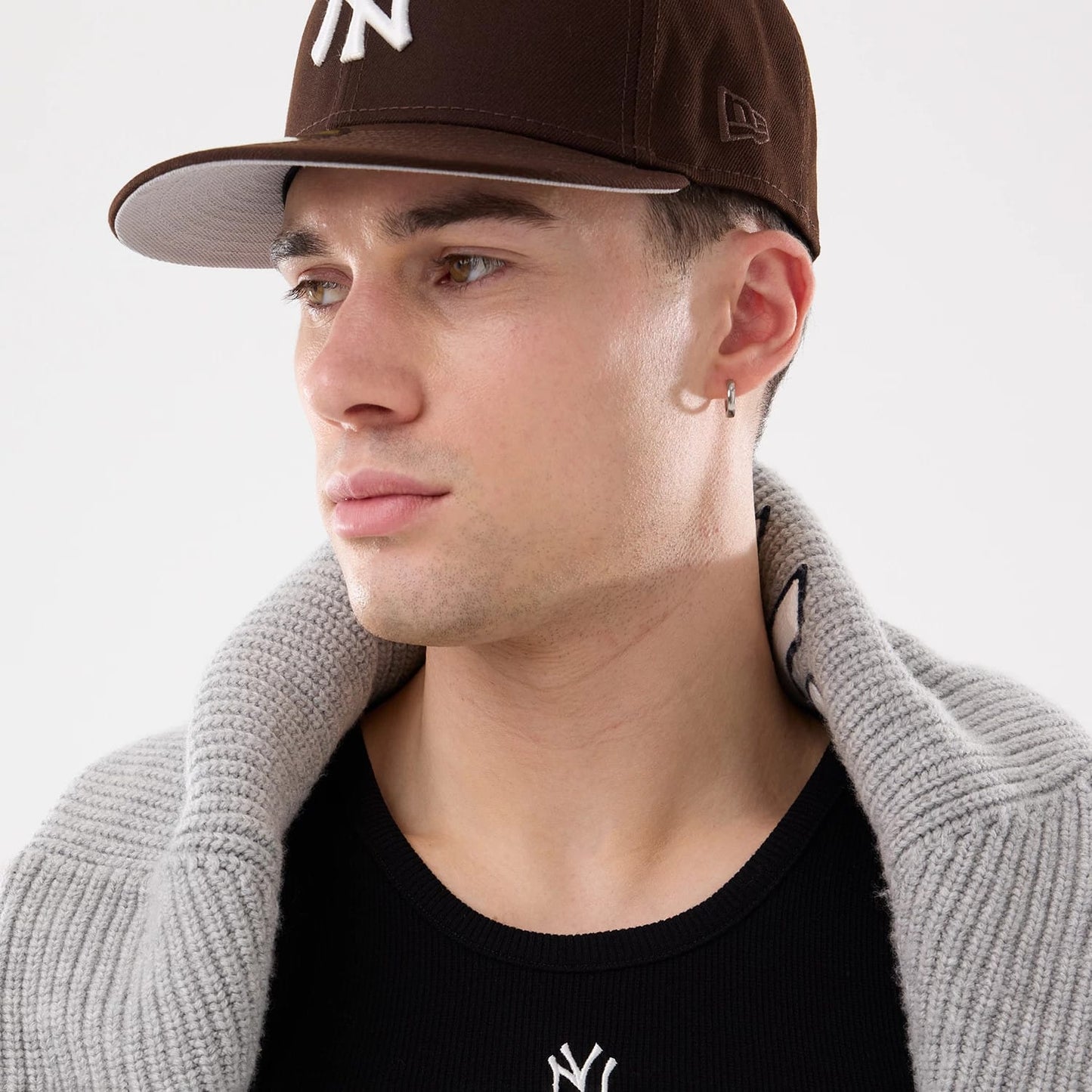 New Era New York Yankees MLB Rib Black Vest, on-model view with a close-up showing the black vest's upper design, featuring a white Yankees logo, alongside a brown cap worn by the model, and a grey knit collar draped over shoulders.