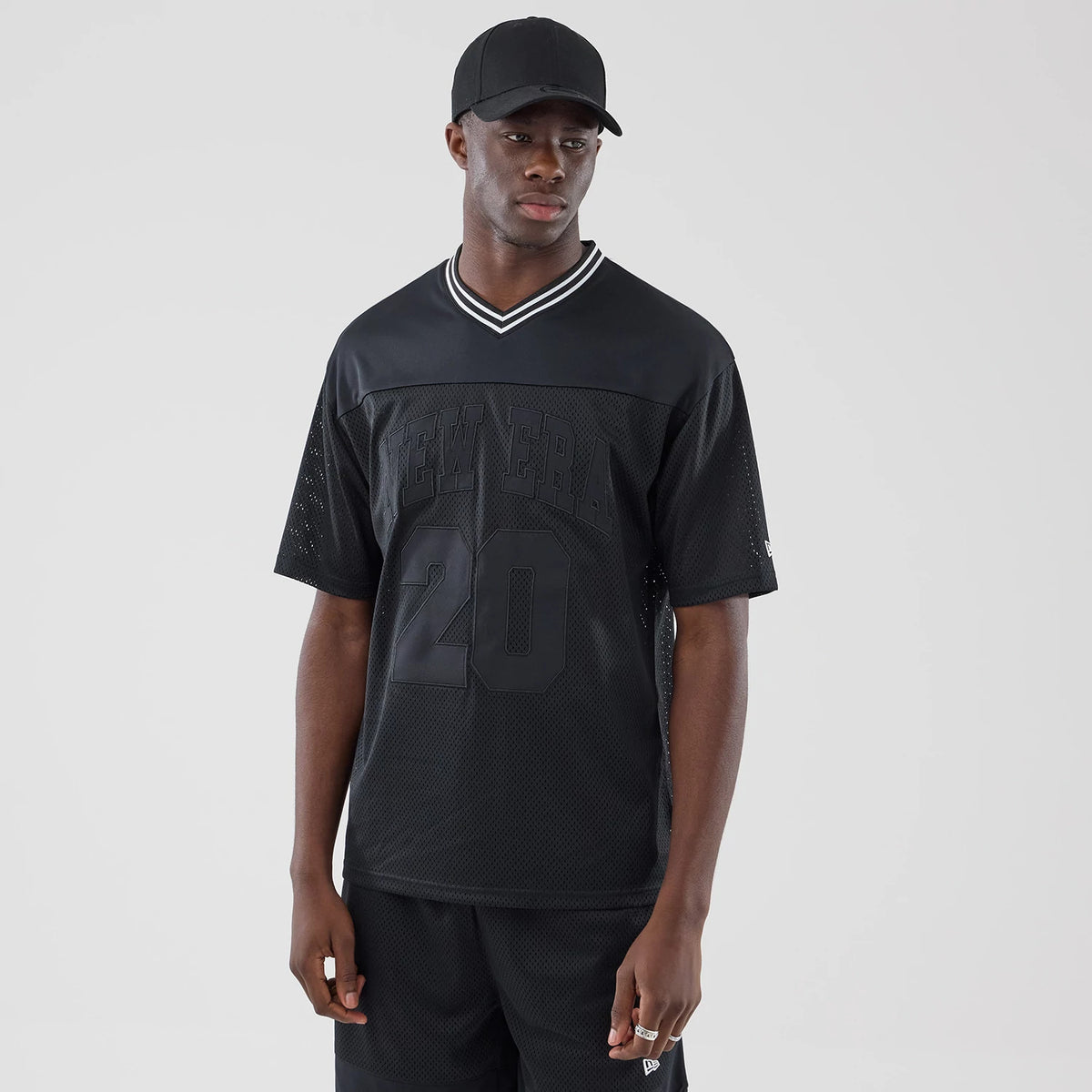 New Era Mesh Black Oversized T-Shirt, front view on-model showing the textured black mesh fabric with large embossed New Era lettering across the chest and the number 20 below, with a contrasting white-striped V-neck collar and short sleeves.