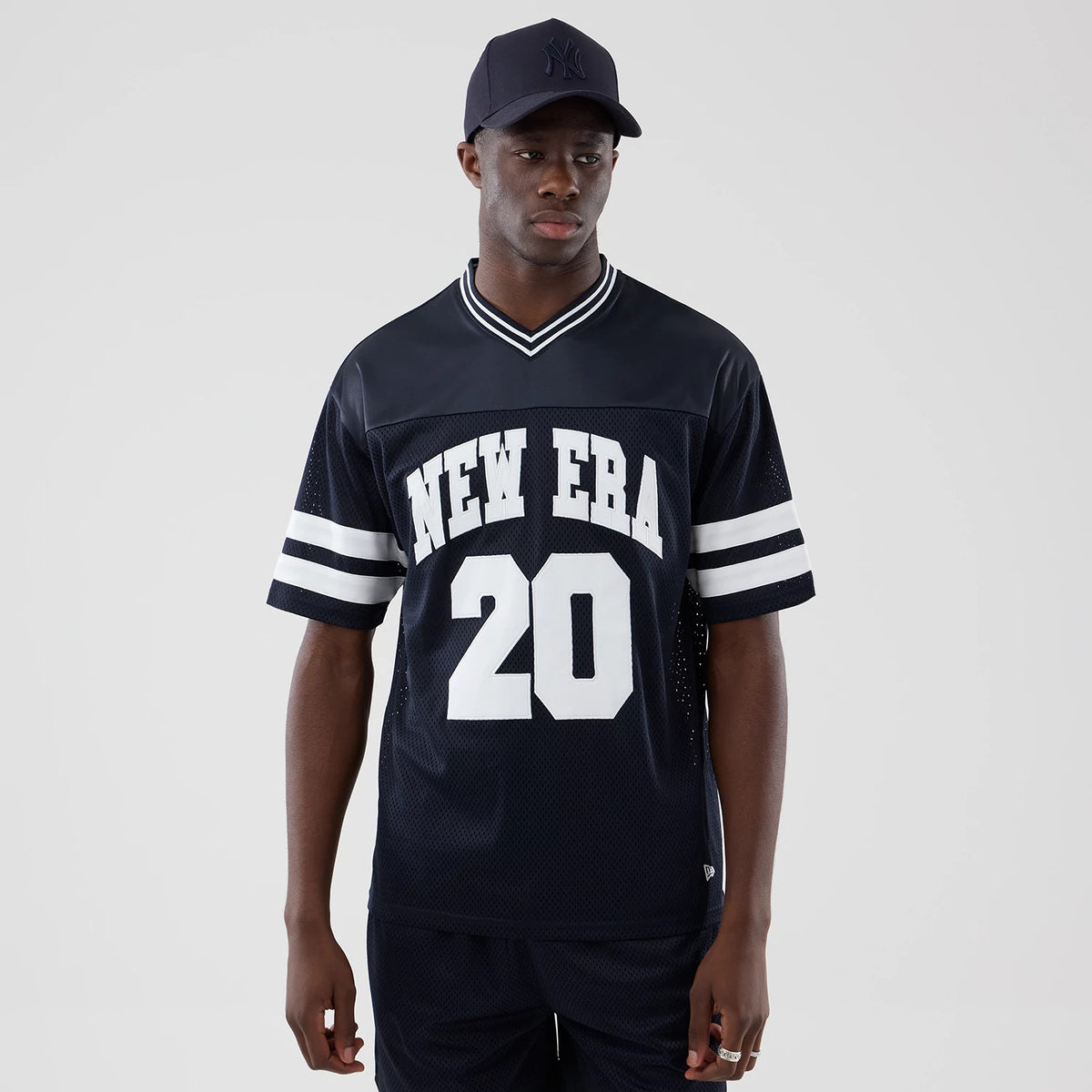 New Era Mesh Navy Oversized T-Shirt, front view on-model showcasing white NEW ERA 20 lettering across the chest, dual horizontal white stripes on the short sleeves, and a contrasting white V-neck ribbed collar.