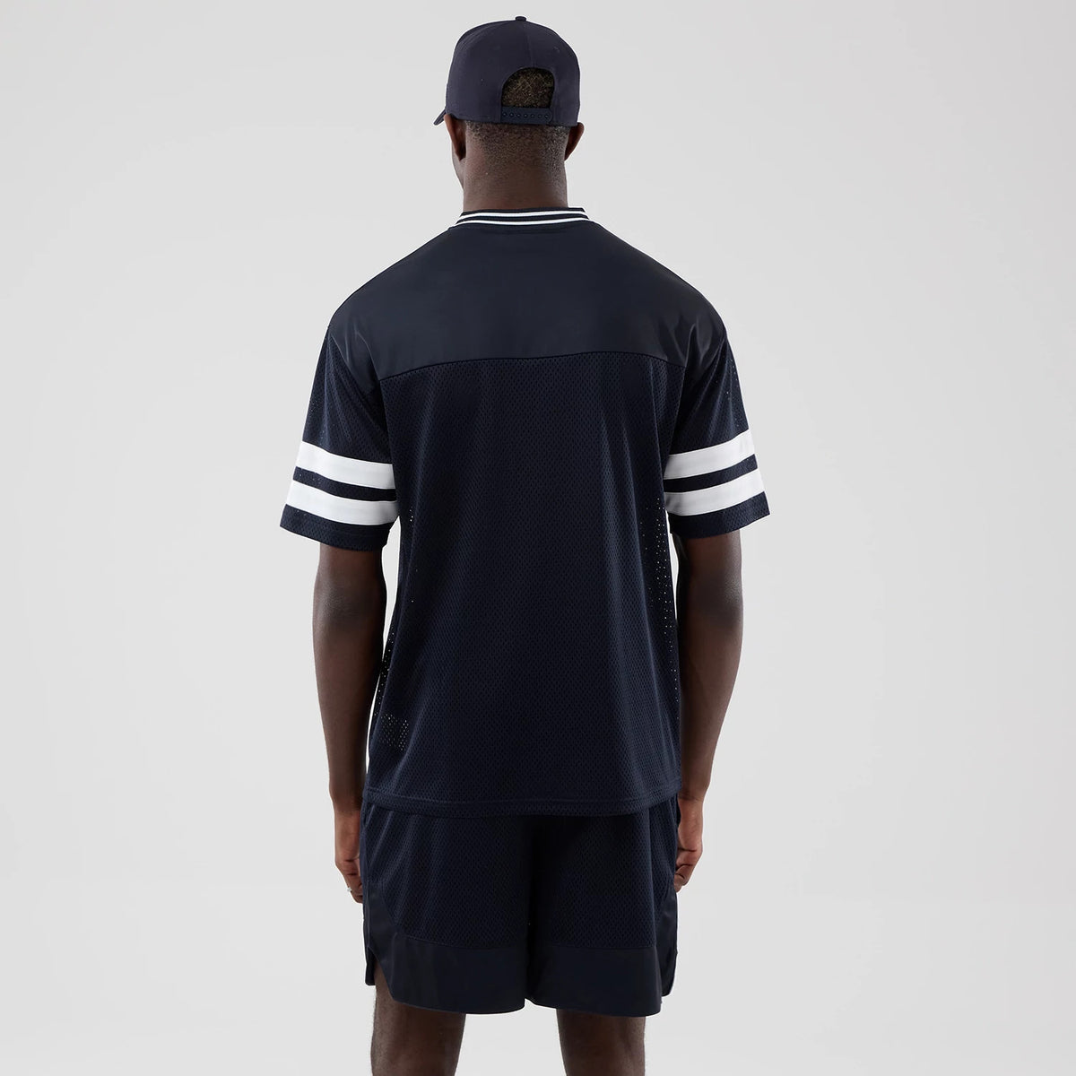 New Era Mesh Navy Oversized T-Shirt, back view on a model showing a mesh texture and contrasting white striped bands on the sleeves, with a dark collar trim, styled with dark shorts and a matching navy cap.