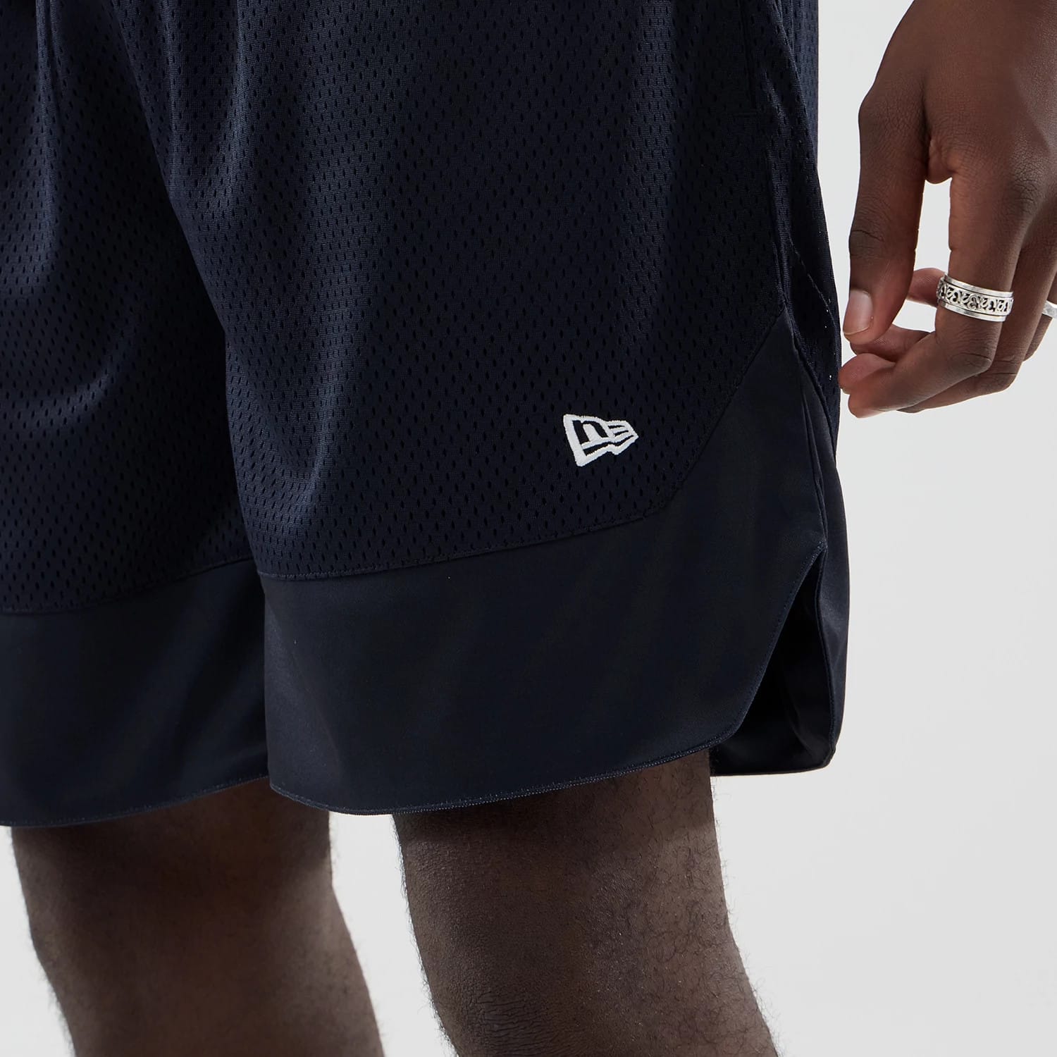 New Era Mesh Navy Oversized Shorts, front view showing the navy mesh fabric with a white embroidered New Era logo on the lower left leg, a black trim at the hem, and a relaxed fit styling on a model's leg.